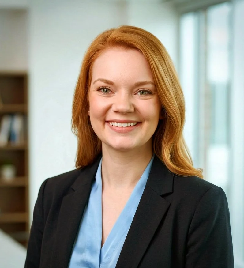A professional woman with red hair smiling, wearing a black blazer and light blue blouse, in an office setting with large windows.