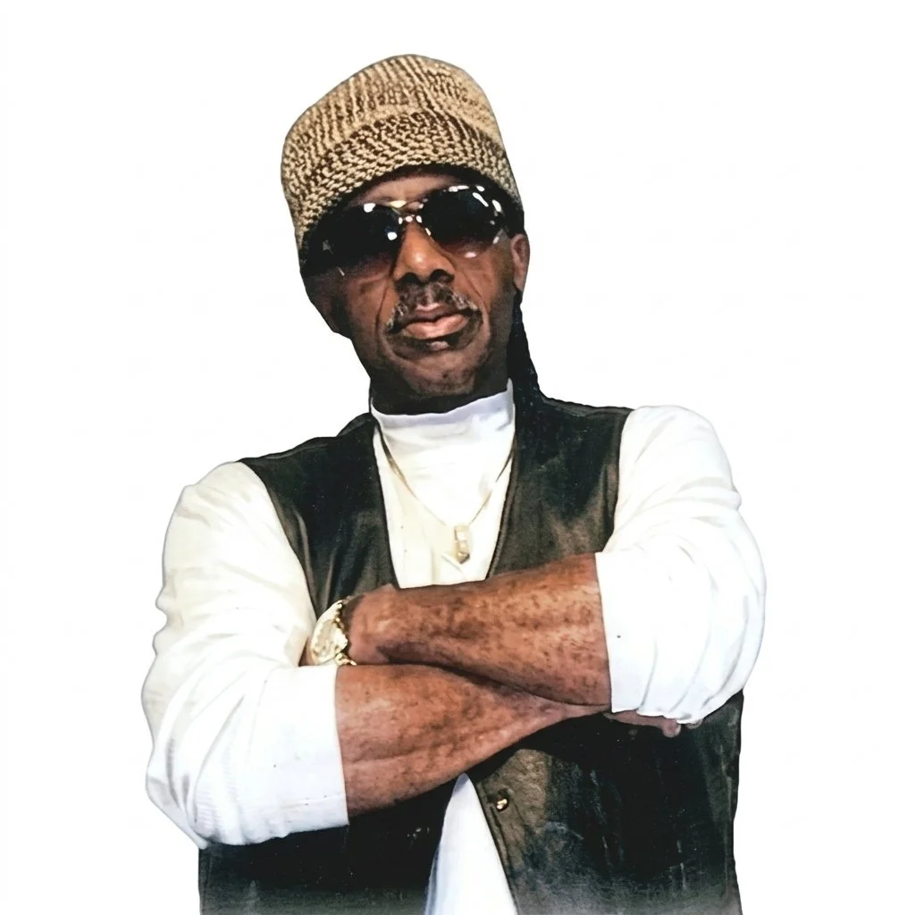 A man wearing sunglasses, a knit cap, a white shirt, and a black vest crossing his arms.