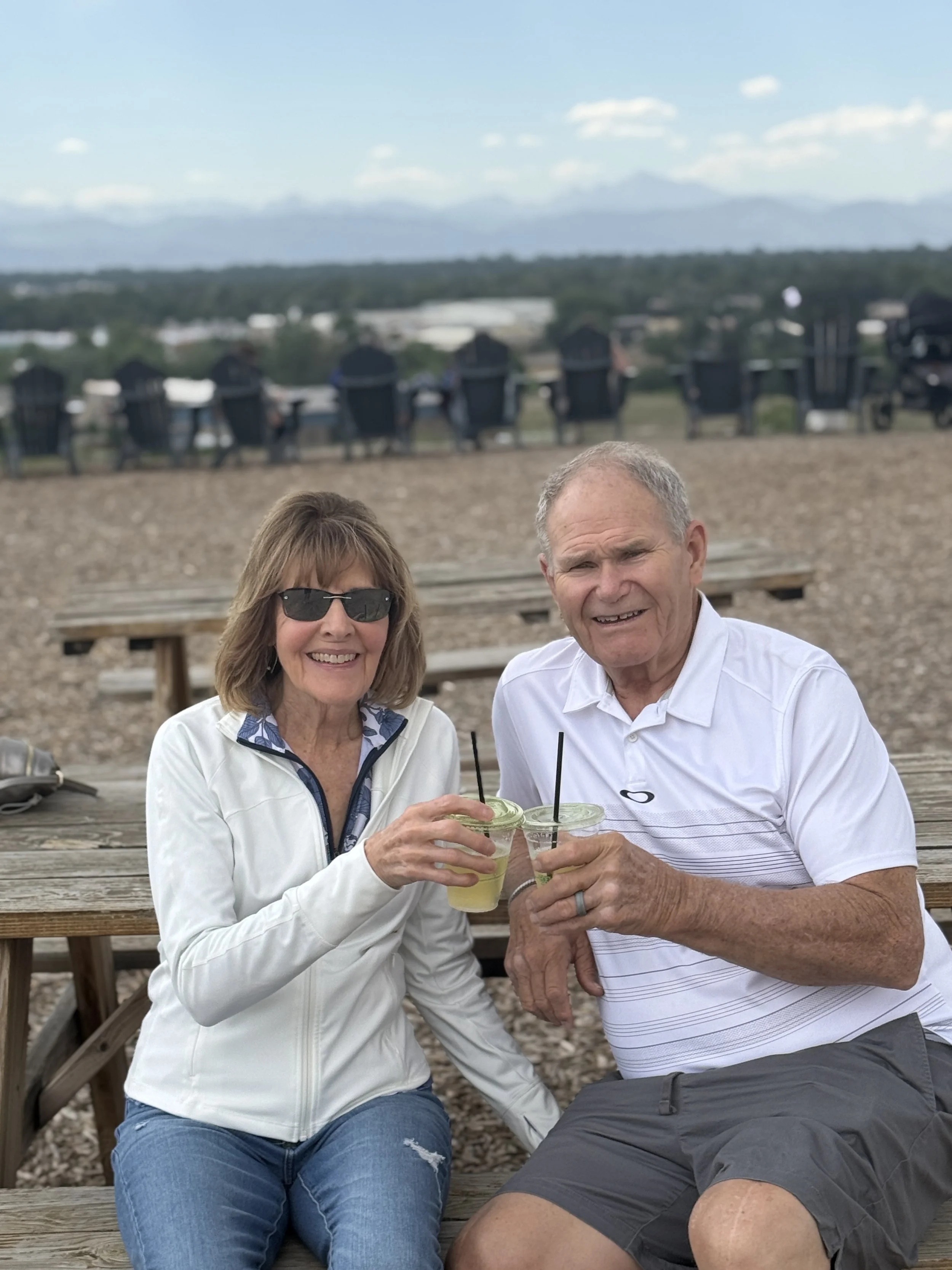 Travis's parents toasting outdoors at a wooden table with a scenic mountain view in the background.