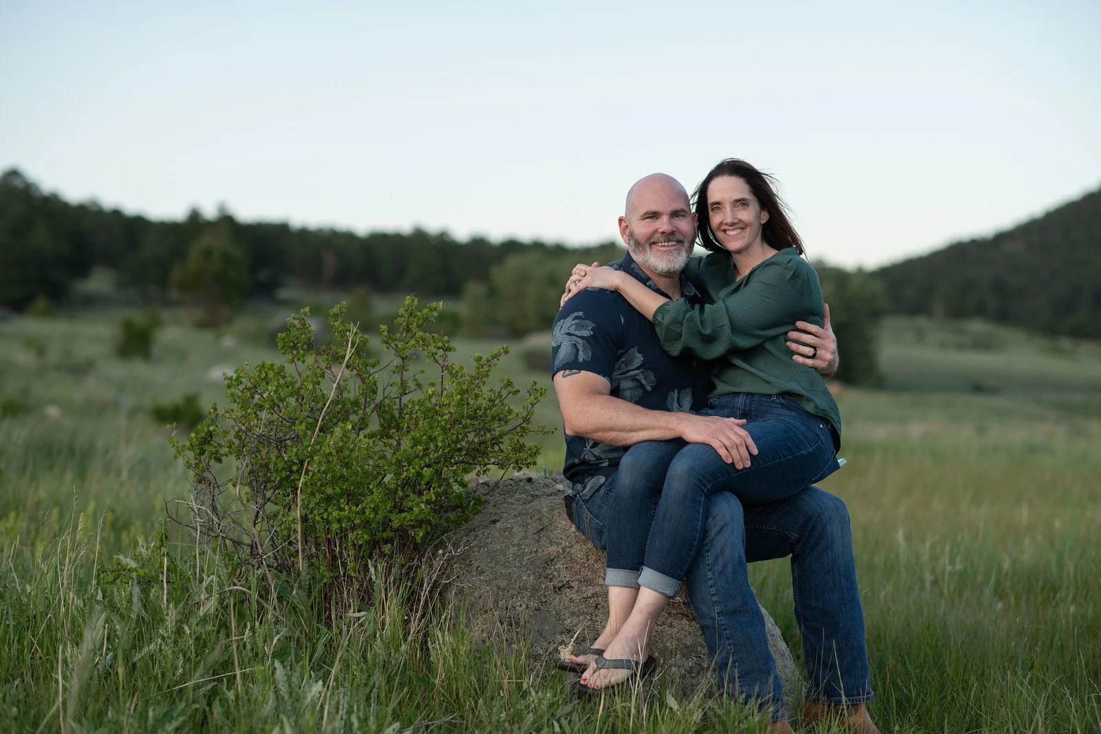A smiling couple sitting on a rock in a grassy field, hugging, with trees and a clear sky in the background.
