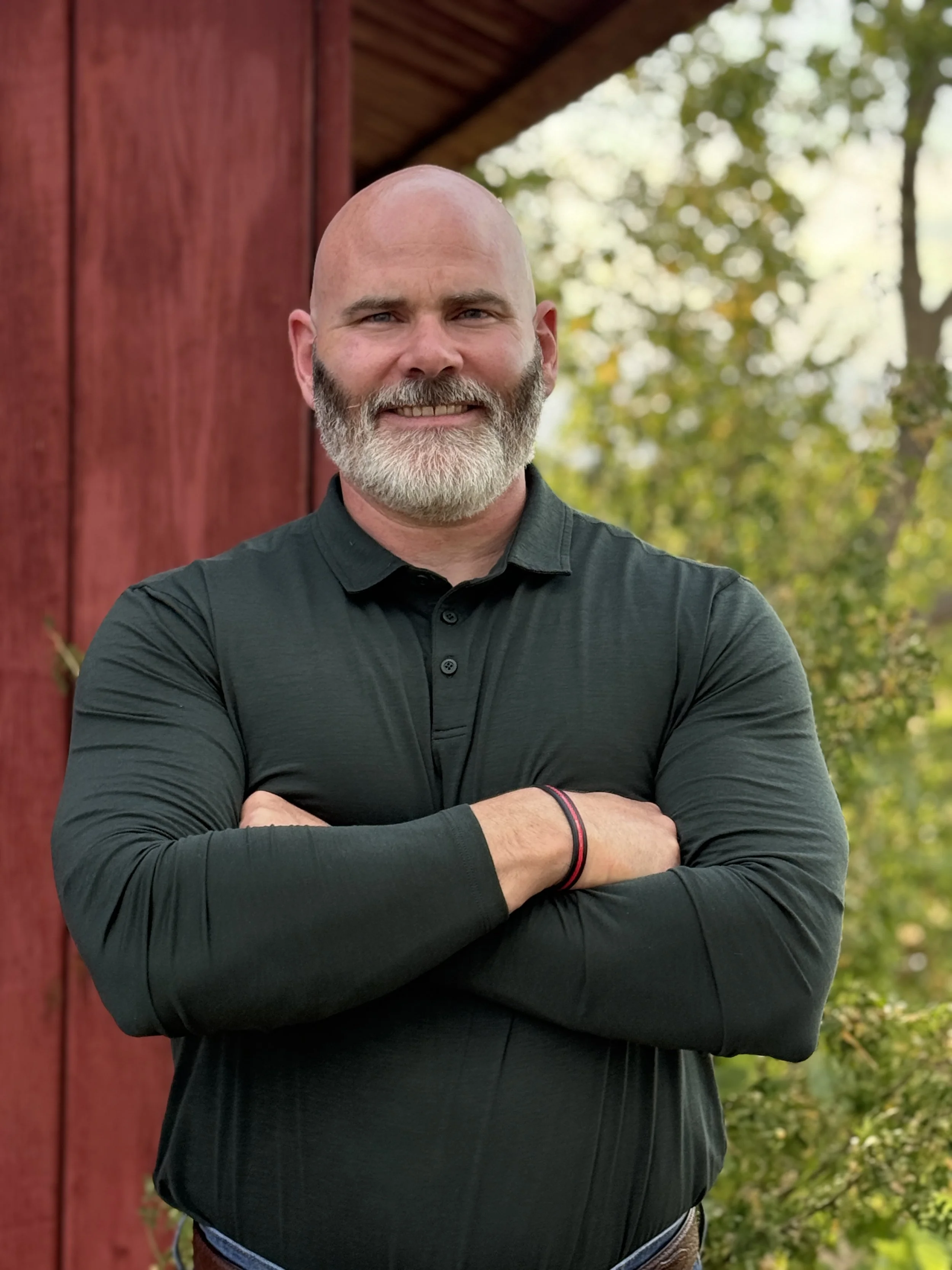 Travis Hovel wearing a dark long-sleeve shirt, crossing his arms in front of a red wooden building and green trees in the background.