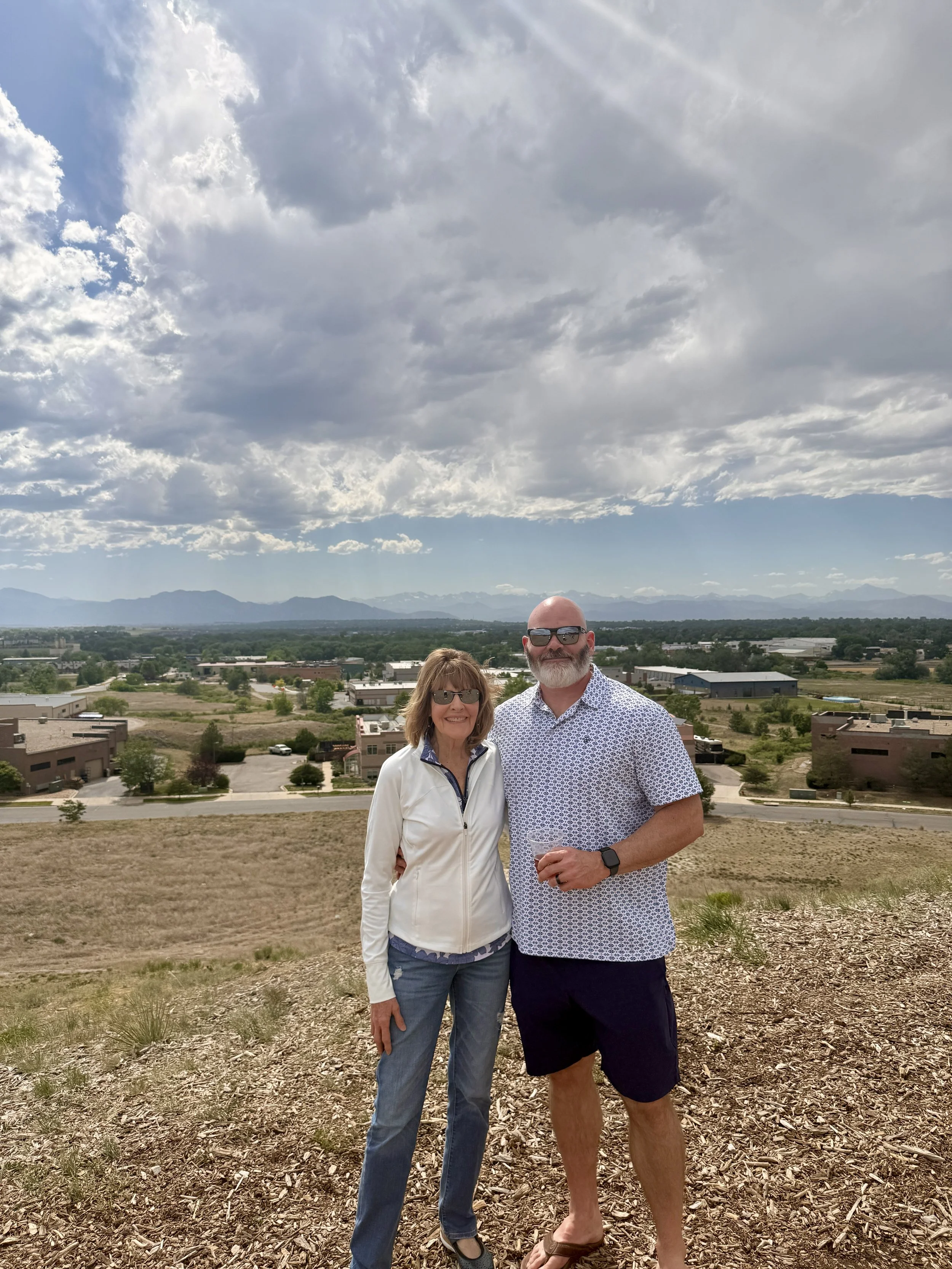 Travis and his mom posing together outdoors on a hillside with a city and mountains in the background, under a cloudy sky.