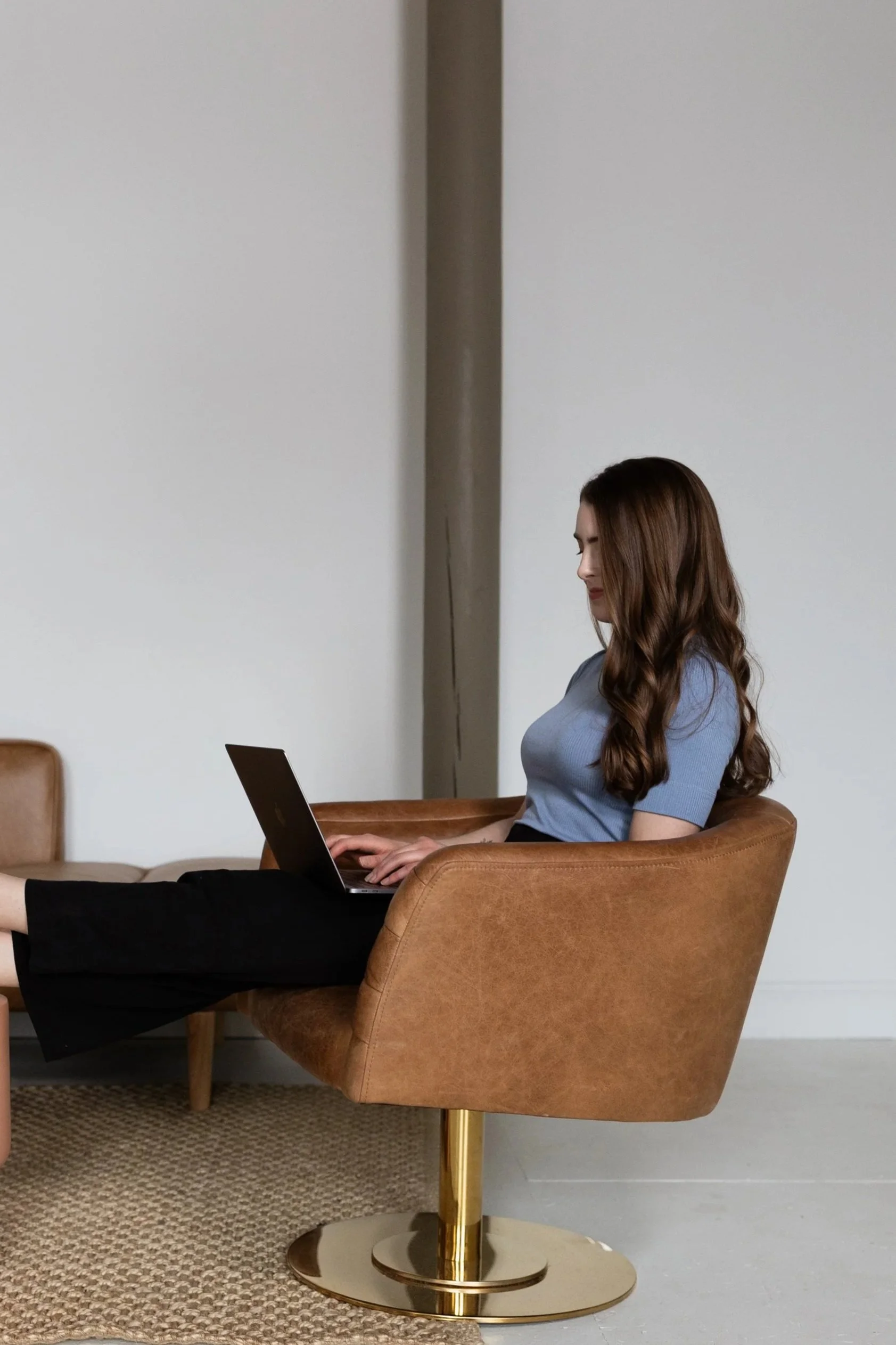 A woman with long brown hair sitting in a tan armchair with a gold base, using a laptop.