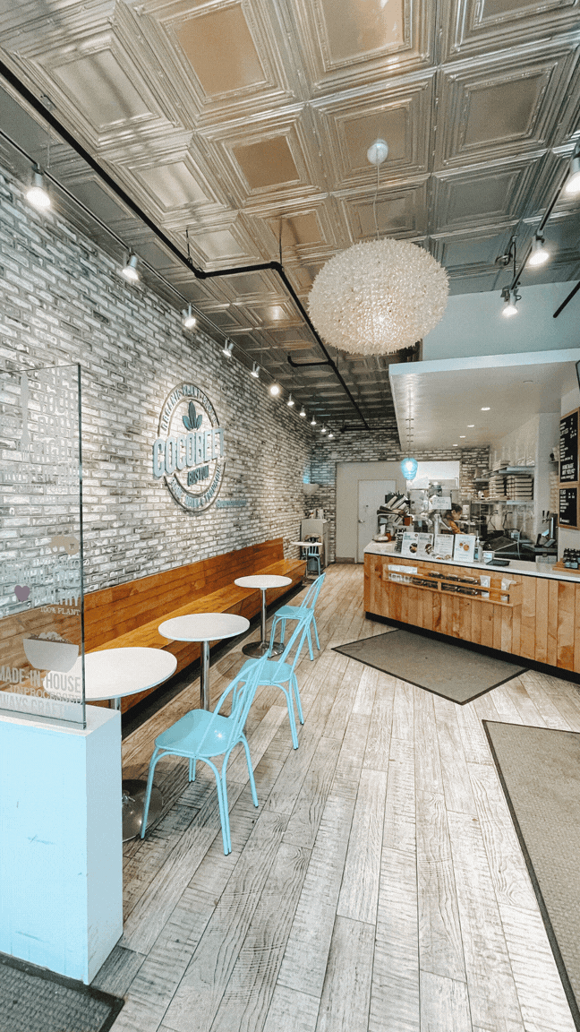 Interior of a modern coffee shop with a brick wall, wooden flooring, white and light blue chairs, small white tables, and a counter area with menu boards and coffee equipment. A large spherical chandelier hangs from the decorative ceiling.