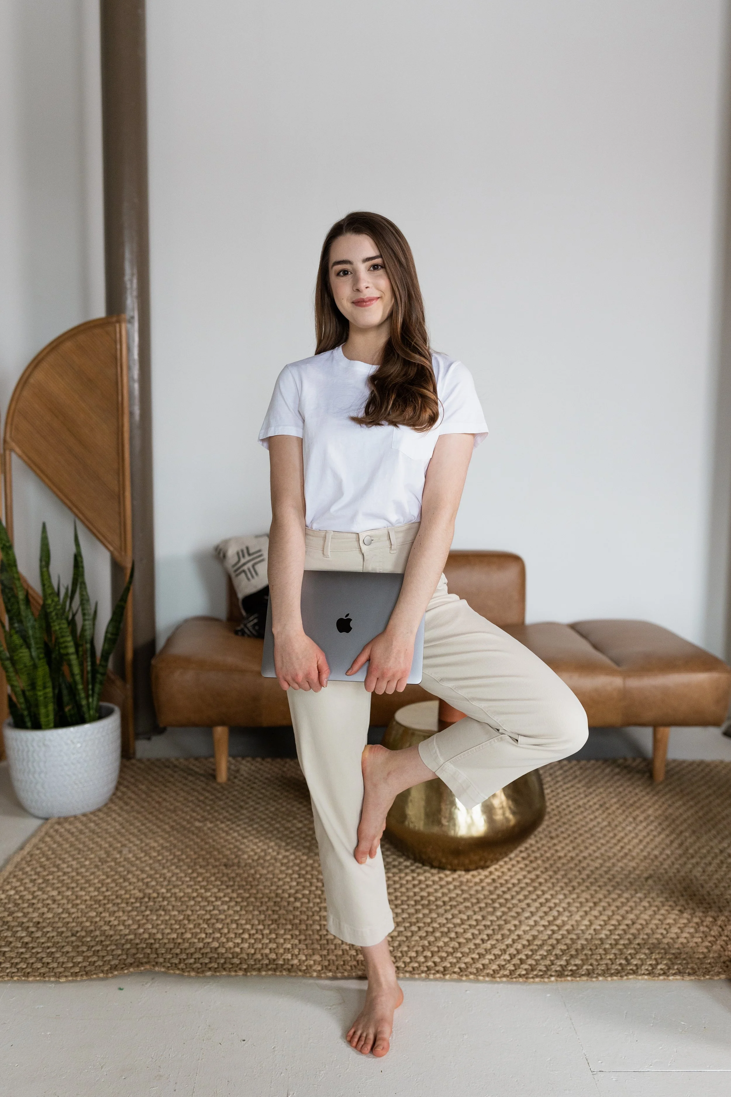 A woman with long brown hair and a white t-shirt standing in a living room, holding a laptop while balancing on one foot. The living room features a brown sofa, a potted plant, and a woven rug.