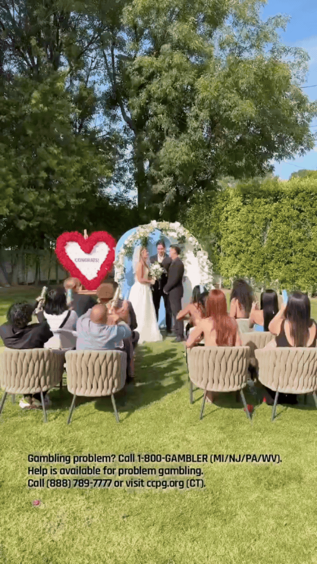 An outdoor wedding ceremony with a bride and groom standing in front of an officiant, under a floral arch, with guests seated in chairs watching. There is a large heart-shaped flower arrangement with the word "CONGRATS!" on it to the left.