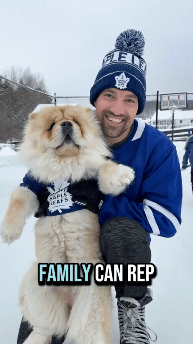 A man wearing a Toronto Maple Leafs hat and blue hockey jersey holding a large, fluffy golden retriever dog in a snowy outdoor setting with a basketball hoop and trees in the background.