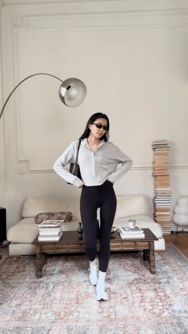 A woman with long dark hair wearing sunglasses, a gray zip-up hoodie, black pants, white sneakers, and a black shoulder bag, standing in a living room with a cream sofa, stacks of books on a wooden coffee table, and a tall stack of books beside the sofa. A modern floor lamp with a metallic rounded shade is overhead.