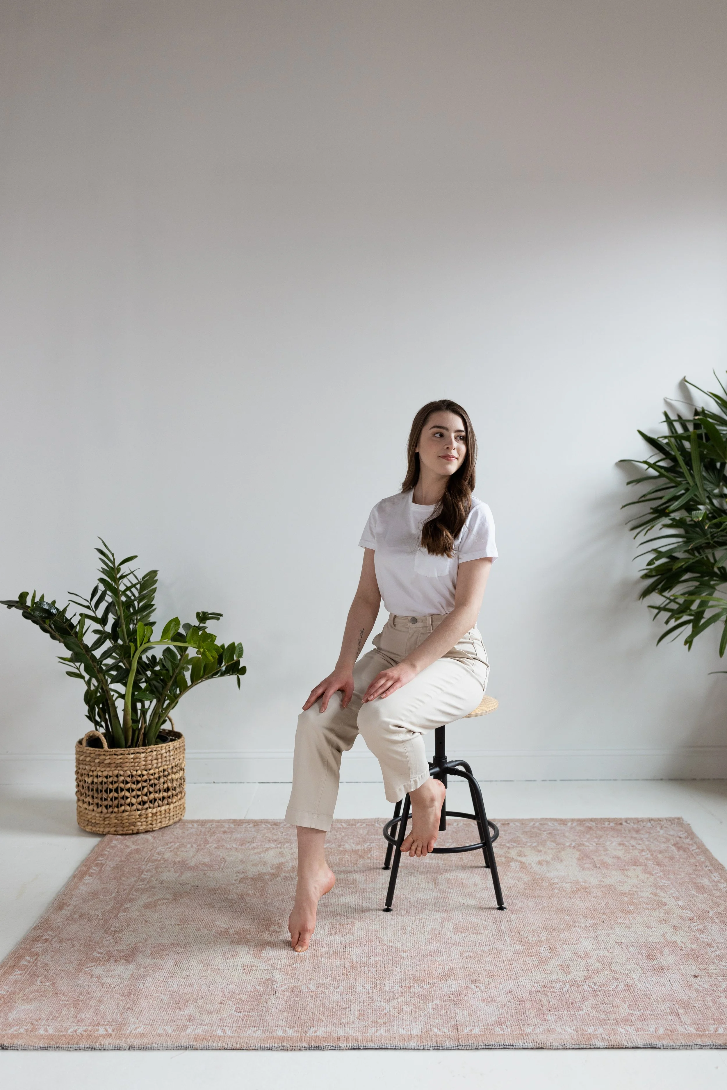 A woman with long brown hair sitting on a black stool in a minimalistic room. She is wearing a white t-shirt and beige pants, with her left hand resting on her lap and her right hand on her knee. She is barefoot, with her left foot touching the floor and her right foot relaxed on the stool. There are green plants in woven baskets on either side of her, and a pinkish beige rug on the light-colored floor.