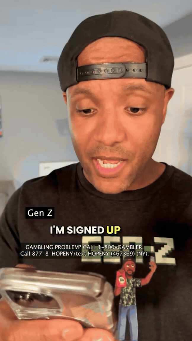 A man wearing a backward black cap and a black T-shirt looking down at his phone, with a blurred background. The T-shirt has text and a graphic of a person raising a fist.