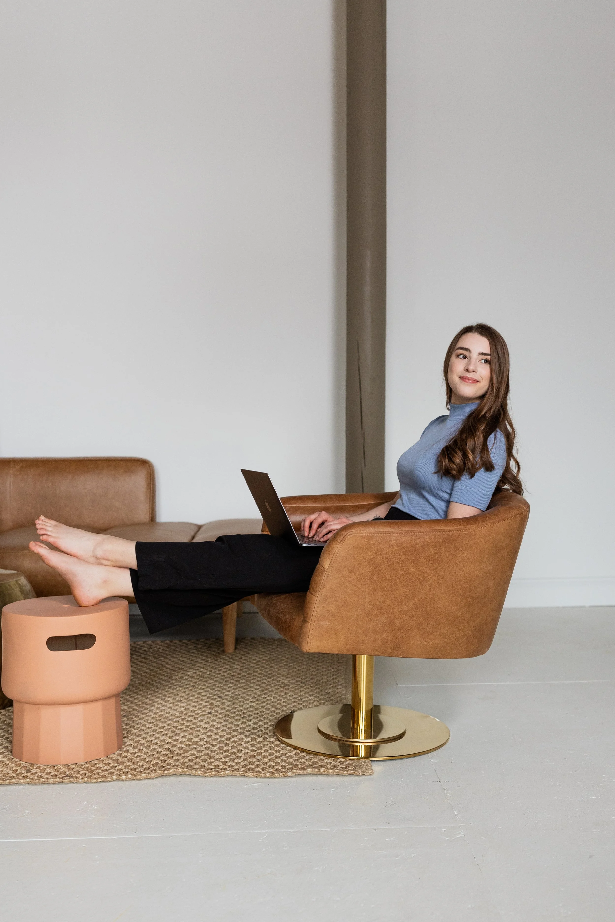 A young woman with long brown hair sits in a brown armchair with her feet resting on a small round pink stool. She is working on a laptop, wearing a blue turtleneck top and black pants, in a modern, minimalistic room with white walls, a textured beige rug, and a tan sofa in the background.