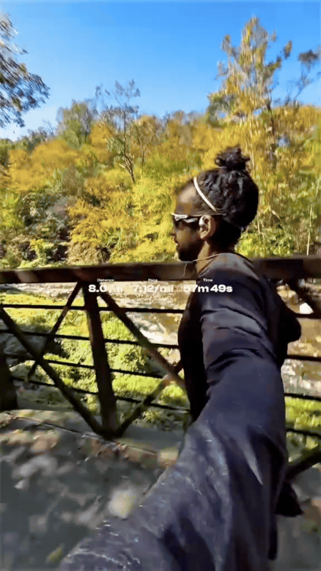 Person taking a selfie on a bridge during a sunny day; trees with green and yellow leaves in the background.