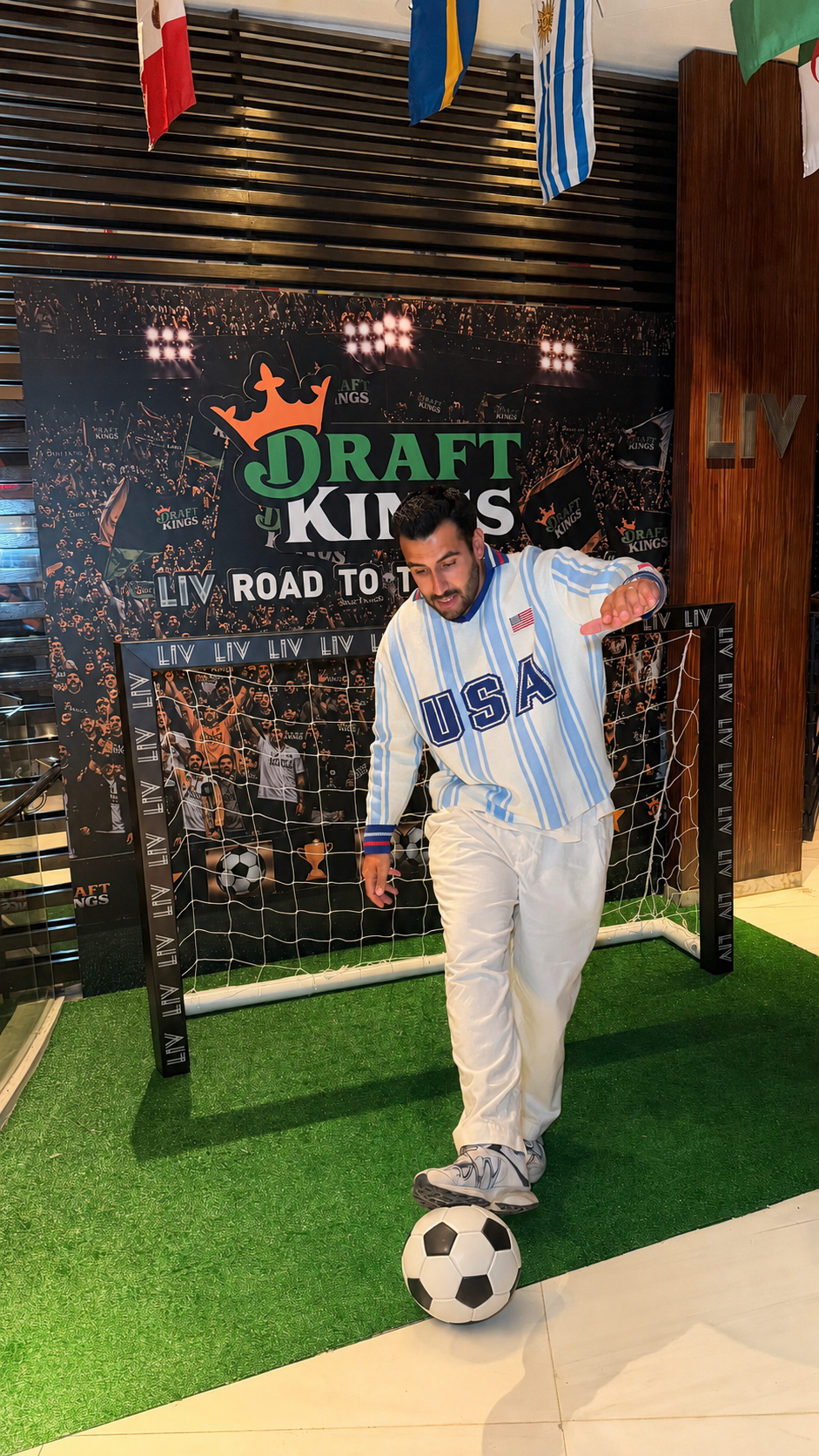 A man in an USA soccer jacket and white pants tries to kick a soccer ball in front of a goal at the Draft Kings event, with a backdrop featuring a crowd and the Draft Kings logo.