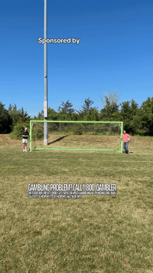 Two men standing on either side of a soccer goal on a grassy field. One is holding a soccer ball. Behind them is a baseball field light pole, trees, and a clear blue sky. Overlaid text in the sky reads "Sponsored by" and lower down, a message about gambling problem help lines.