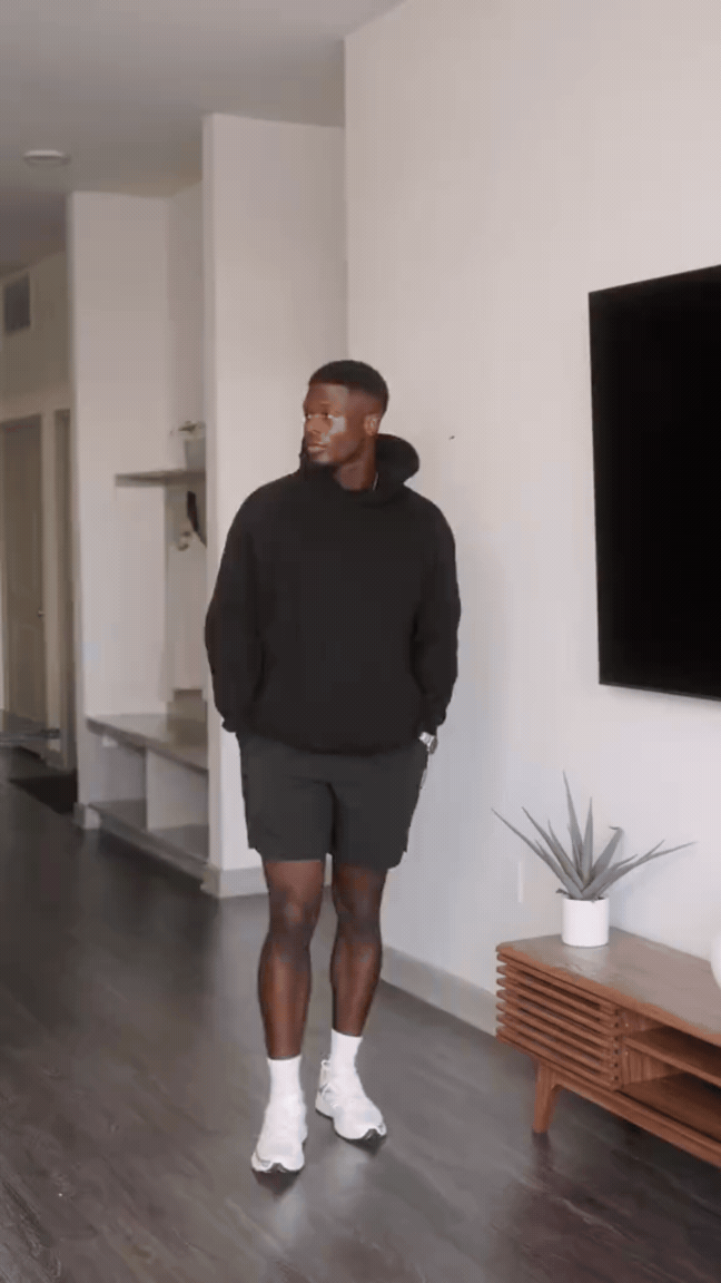 A man standing in a modern apartment with hardwood floors, wearing a black hoodie, black shorts, white sneakers, and white socks, with a white potted plant on a wooden table nearby.