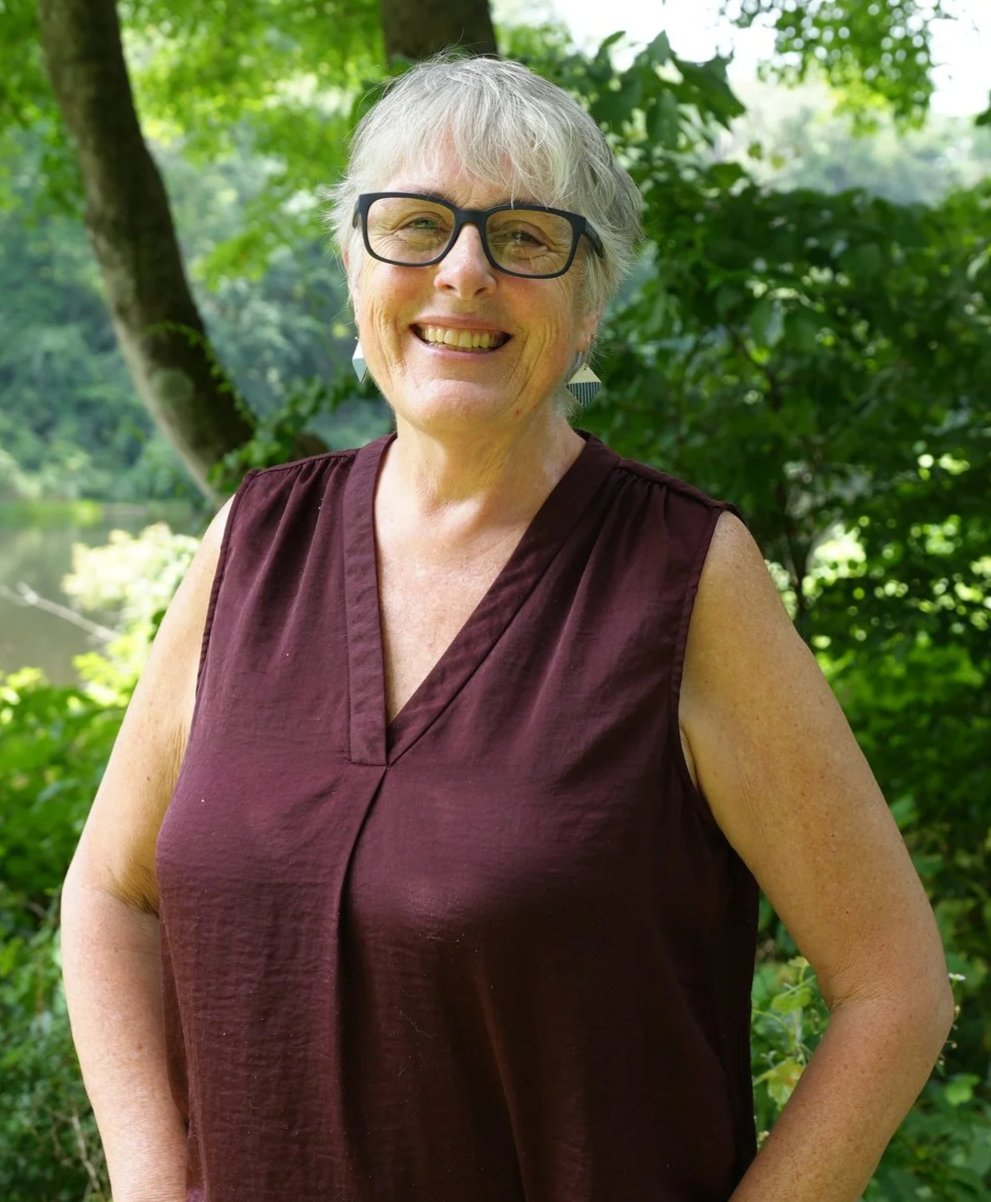 A smiling older woman with short gray hair and glasses standing outdoors in a lush green park with trees and a body of water in the background.