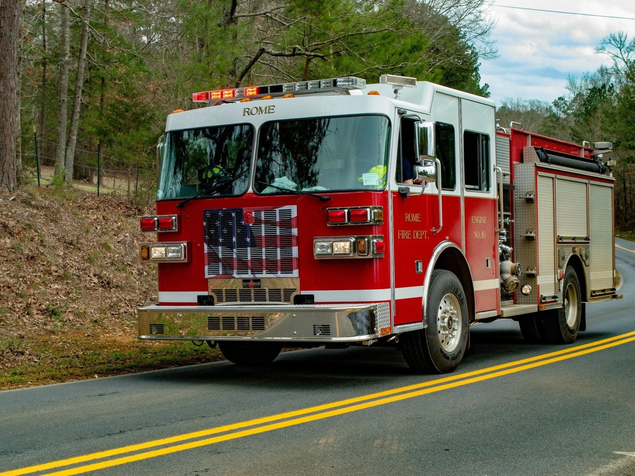 A red fire truck with white accents labeled 'Rome Fire Dept' is parked on the side of a rural road with double yellow lines. The scene includes trees and a cloudy sky in the background.