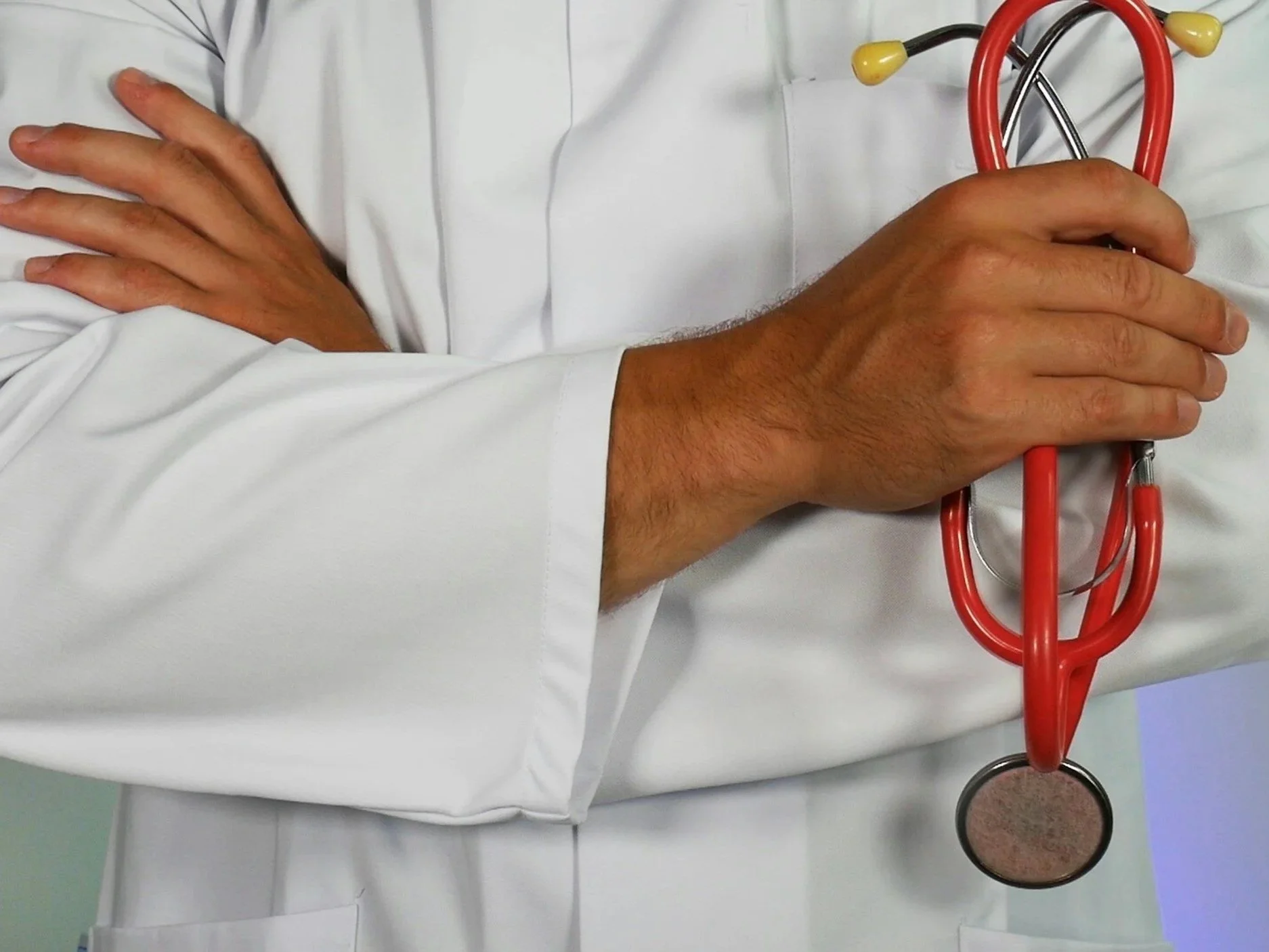 A healthcare professional in a white coat holding a red stethoscope against their chest, with a background of a mint green wall.