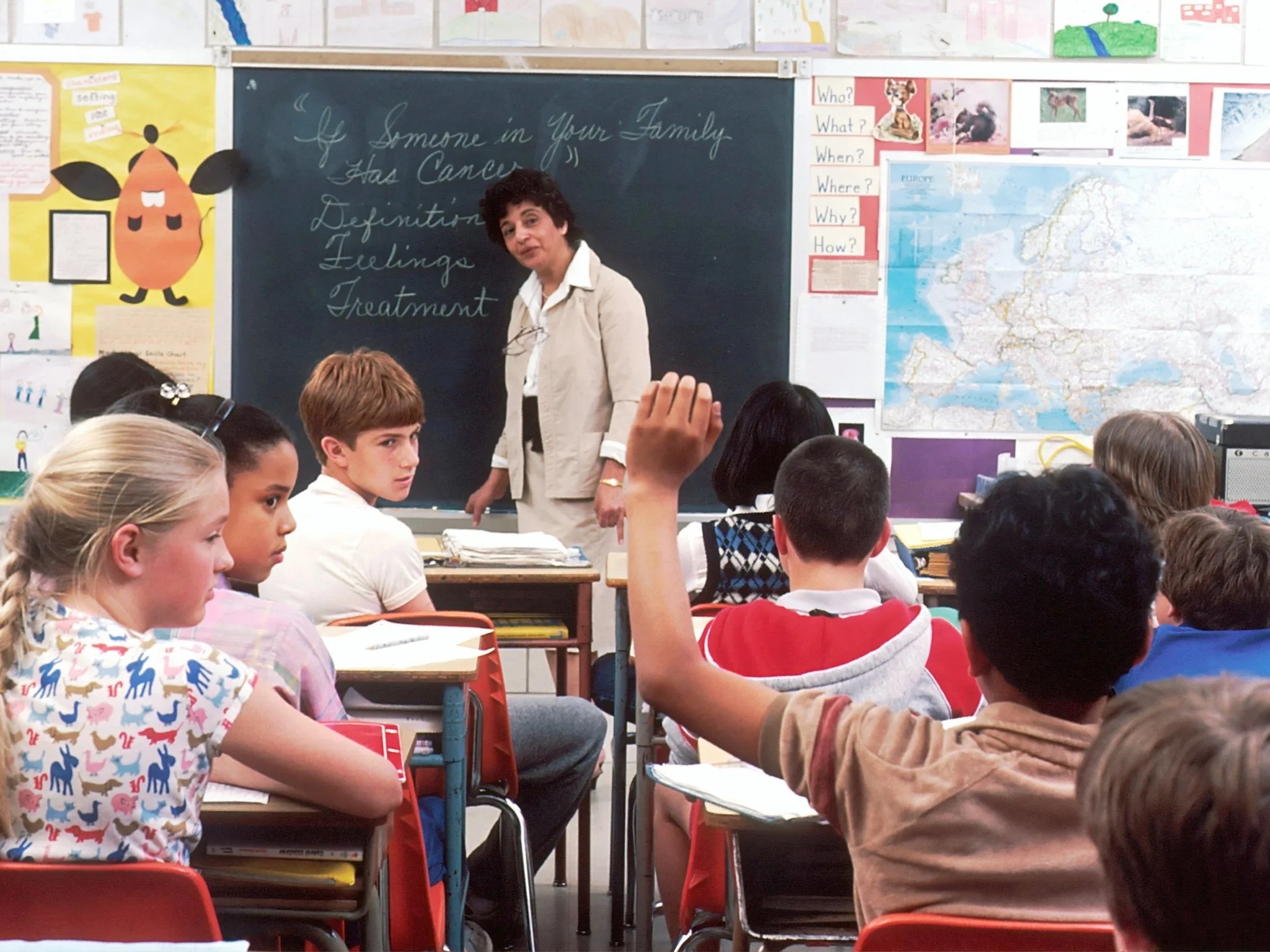 A classroom with students sitting at desks, raising their hands, and a female teacher standing in front of a blackboard with writing. The classroom has posters, maps, and children's artwork on the walls.