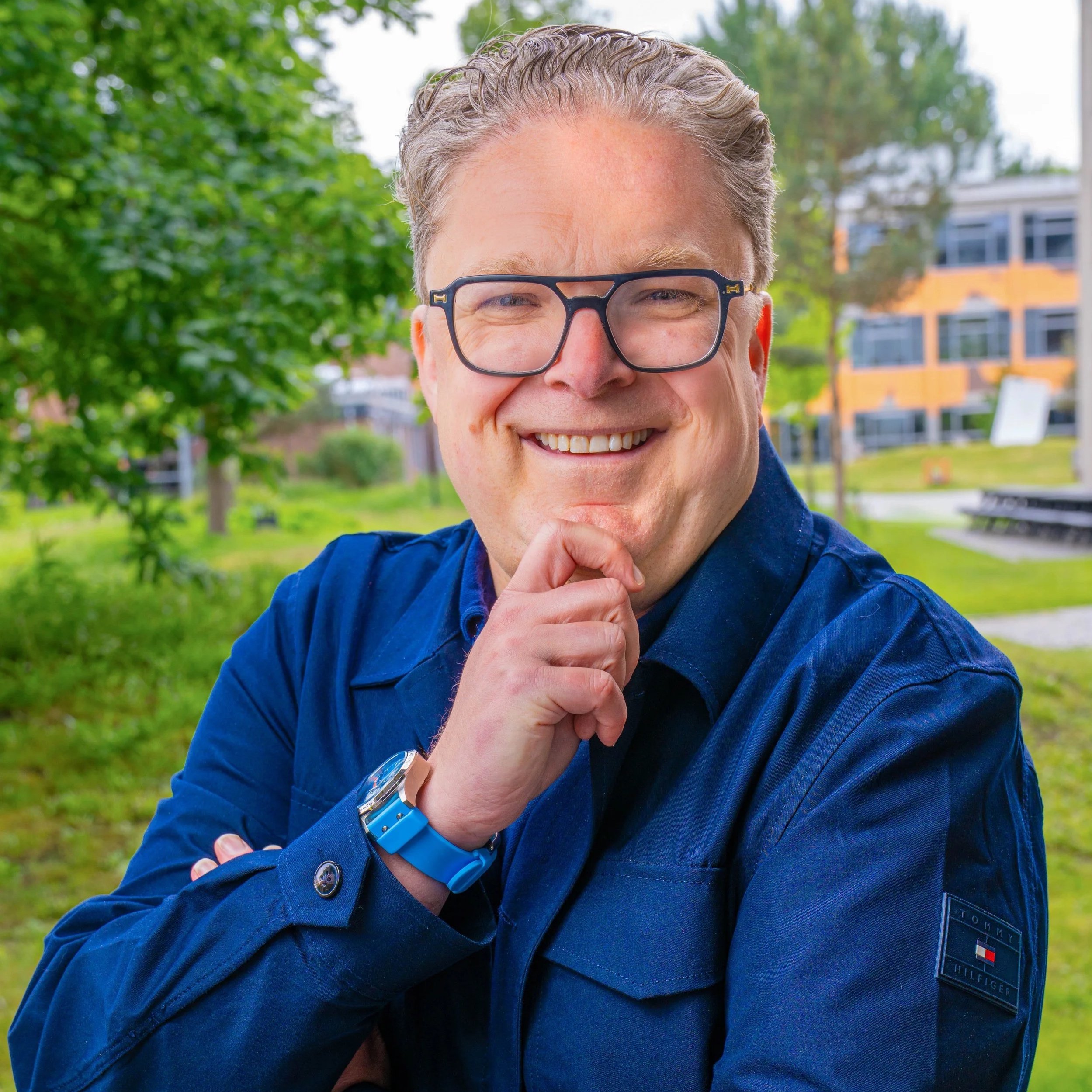 Close-up of a smiling man with glasses, wearing a blue jacket and watch, standing outdoors with green trees and buildings in the background.
