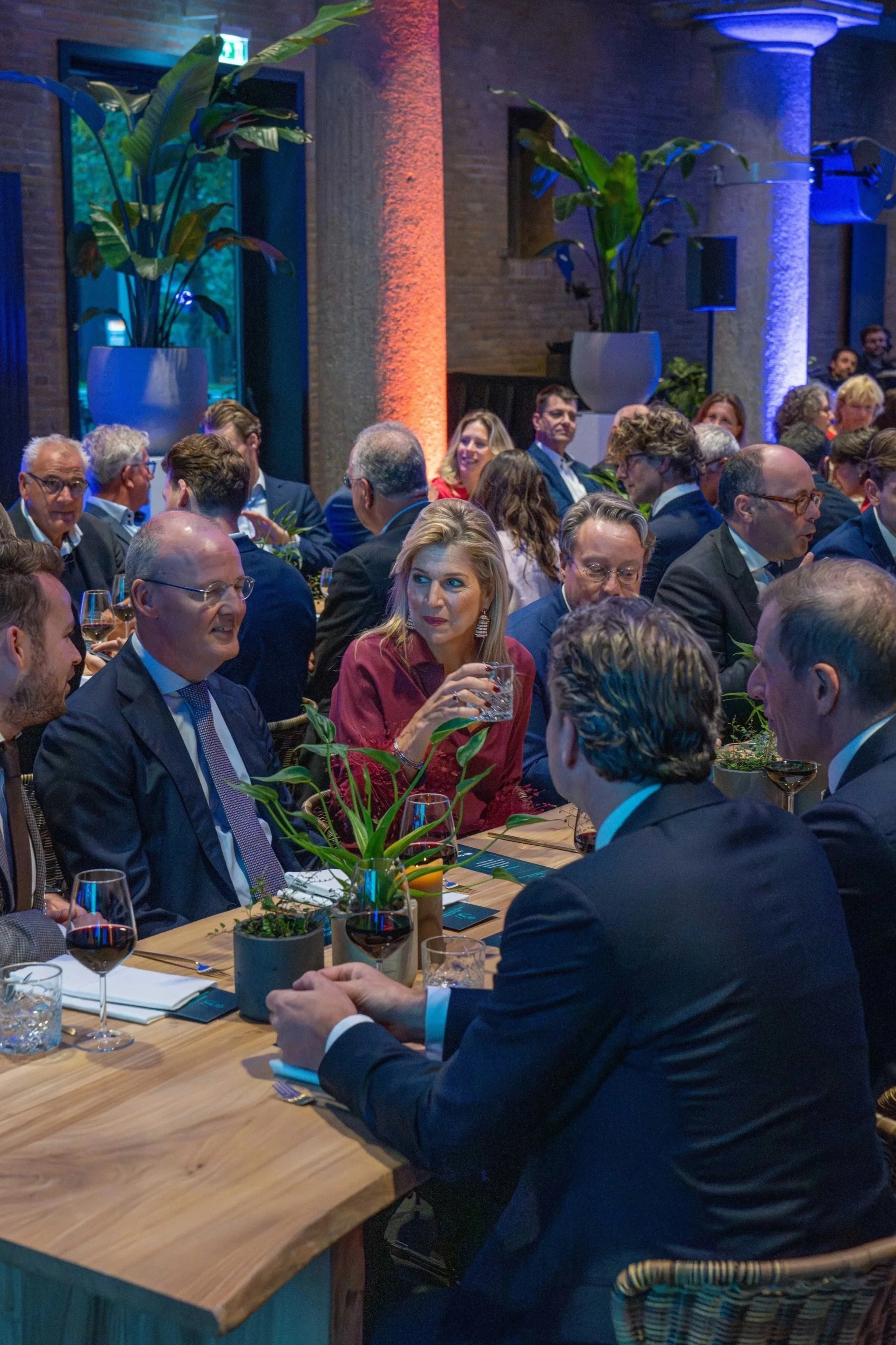 People gathered at a conference or dinner event, seated around a wooden table decorated with plants, glasses of red wine, and small notepads, in a dimly lit room with colorful lighting and large plants.