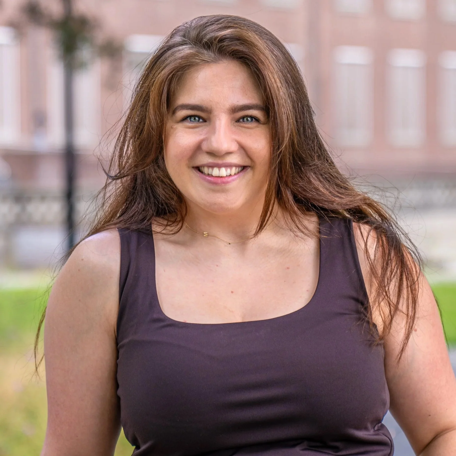 Close-up of a smiling woman with long brown hair, wearing a dark sleeveless top, outdoors in a park setting.