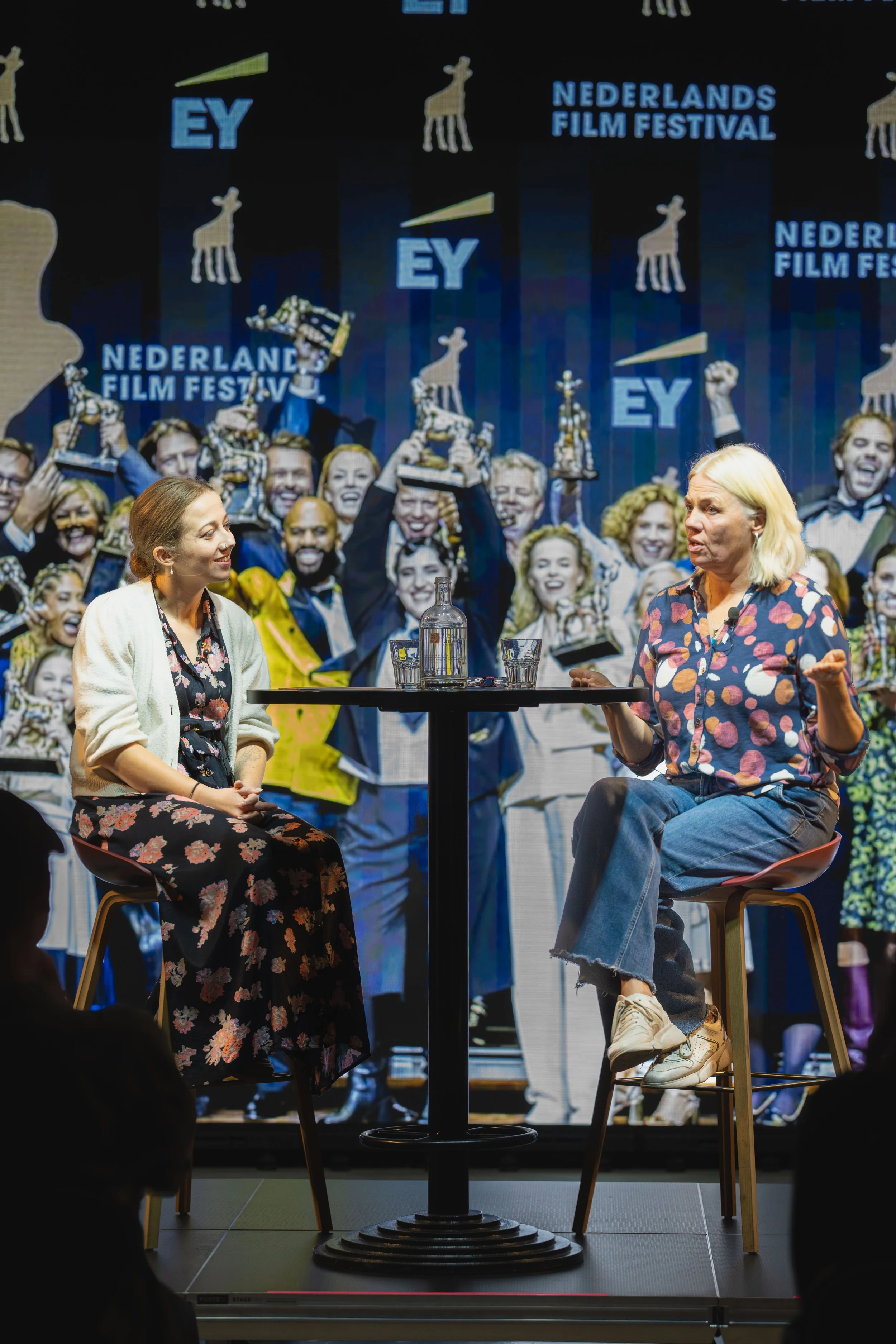 Two women are seated on stage, engaged in a conversation, with a large background screen showing the Netherlands Film Festival logo and a group of people celebrating with trophies, giraffe illustrations, and the EY logo.