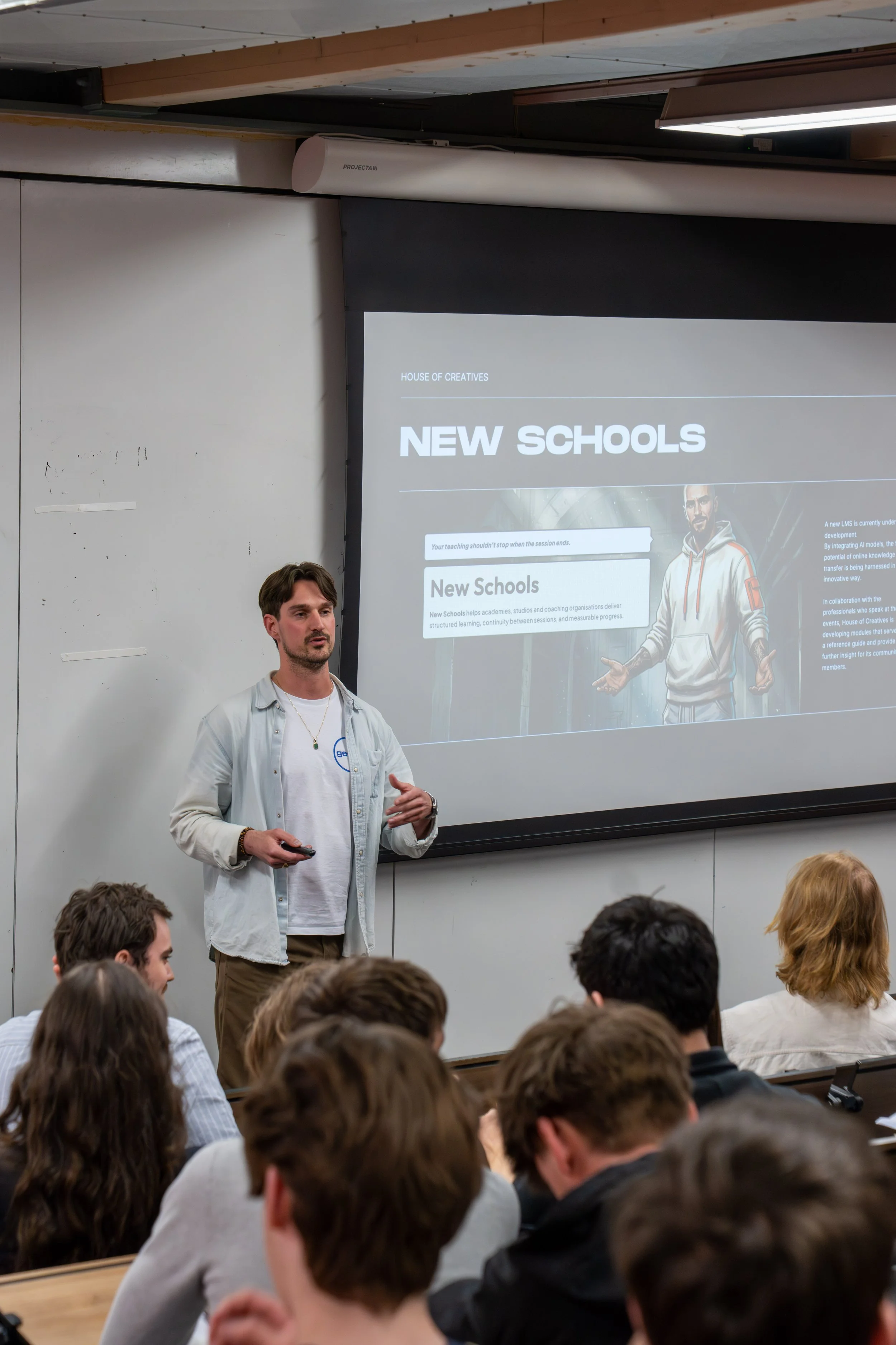 A man with brown hair and a beard is standing at the front of a classroom, giving a presentation to students. He is wearing a white shirt over a white t-shirt and is holding a remote control. Behind him, there is a large screen displaying a slide titled 'New Schools' with images and text about innovative education concepts. Students are seated and listening attentively.