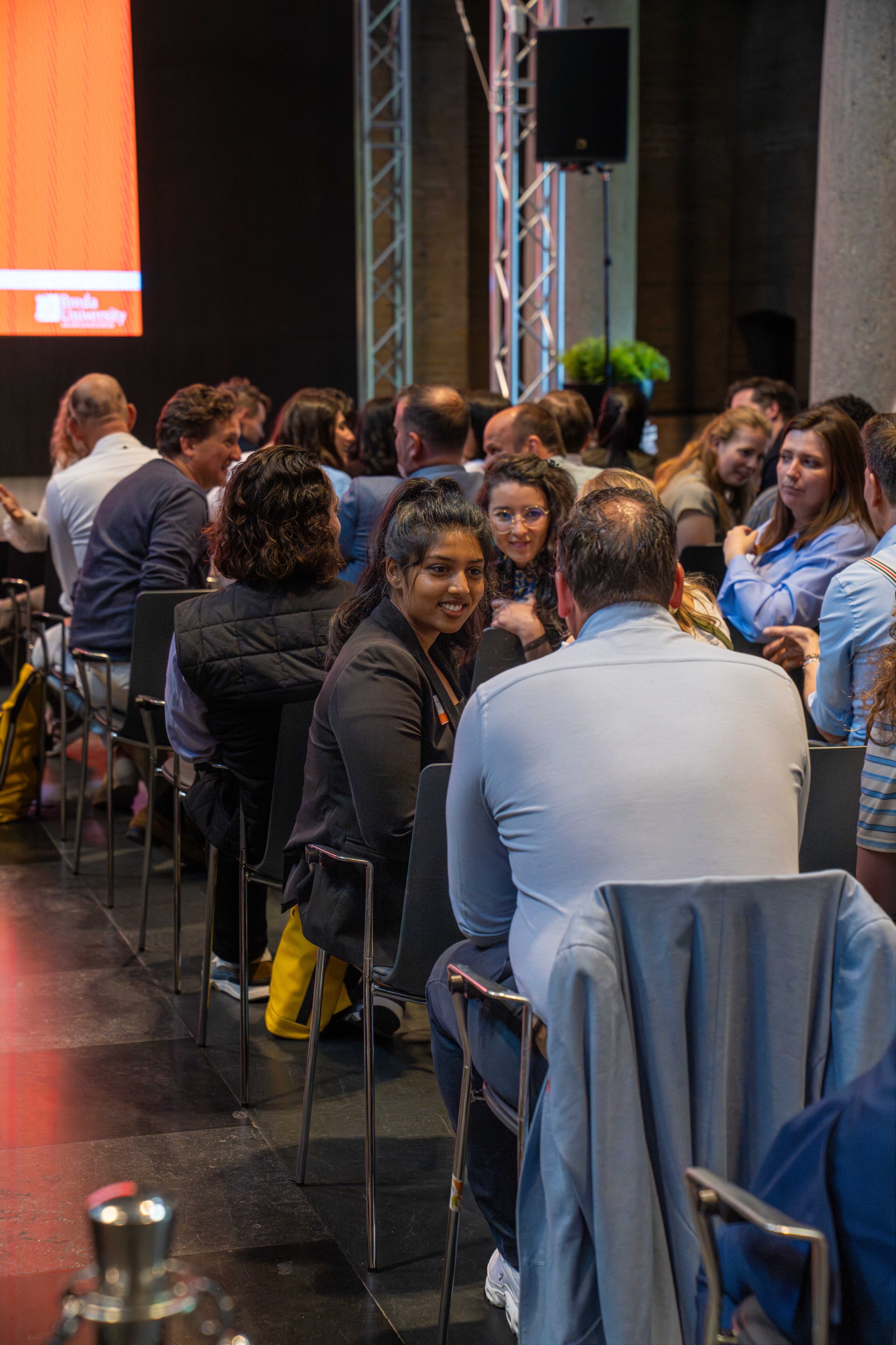 People sitting at a conference or seminar, facing toward the front with a large screen and stage setup, engaged in conversation or listening.