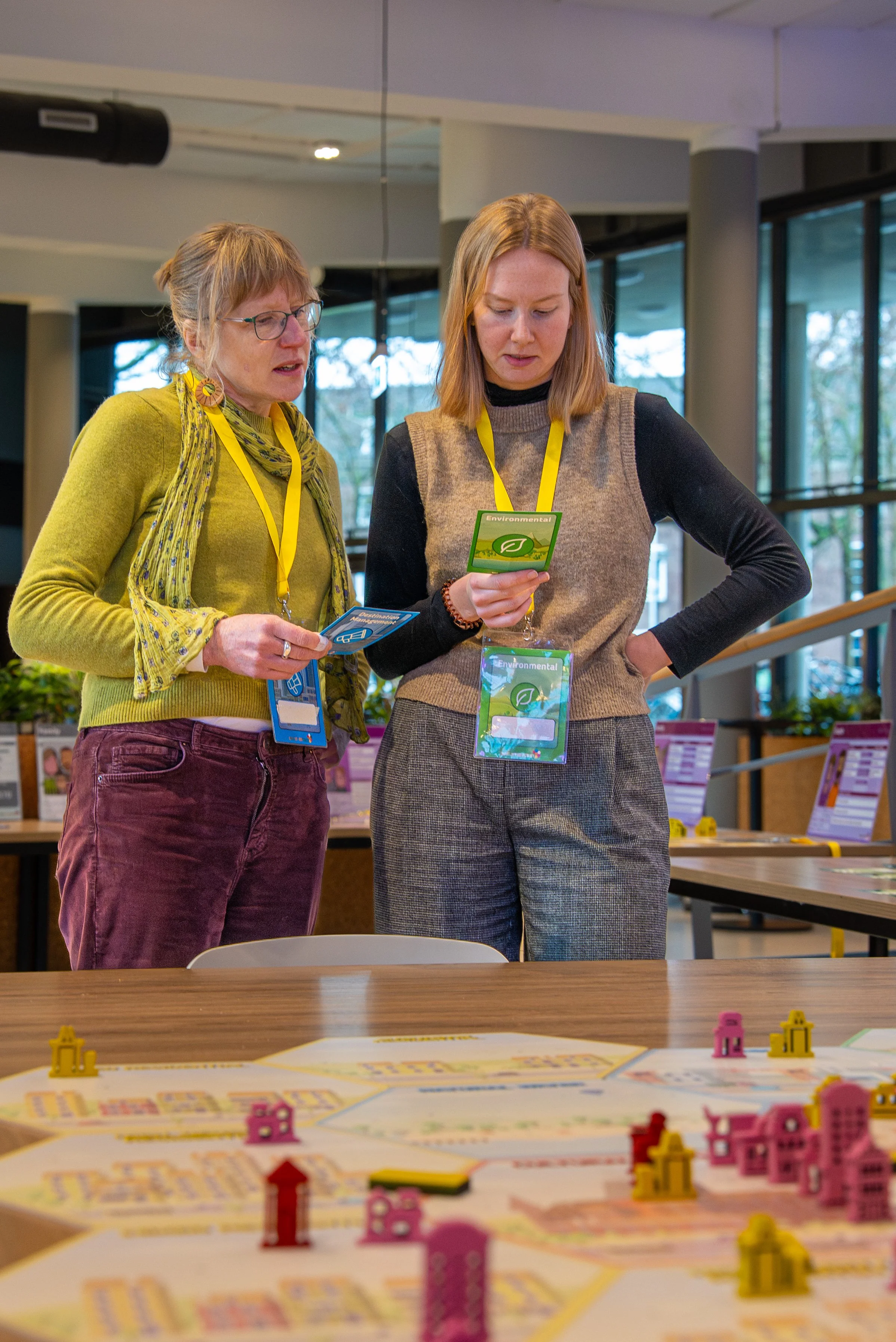 Two women are standing inside a room, looking at a card or booklet. One woman has glasses and a yellow cardigan with a scarf, while the other has blonde hair and is wearing a tan vest over a black long-sleeve shirt. They both have yellow lanyards around their necks.