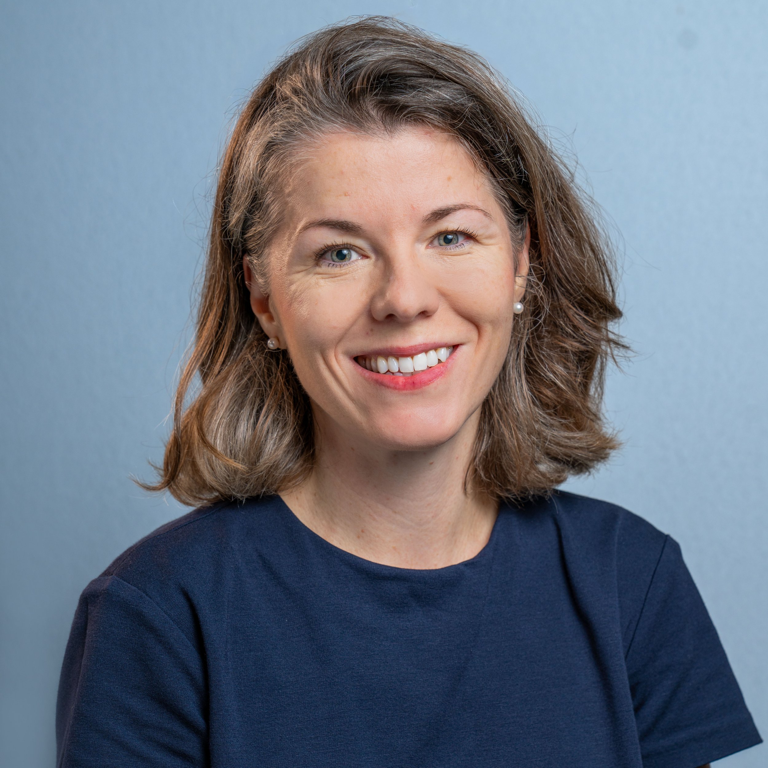 A close-up of a woman's face and upper body, with shoulder-length wavy brown hair, wearing pearl earrings and a navy blue top, smiling against a light blue background.
