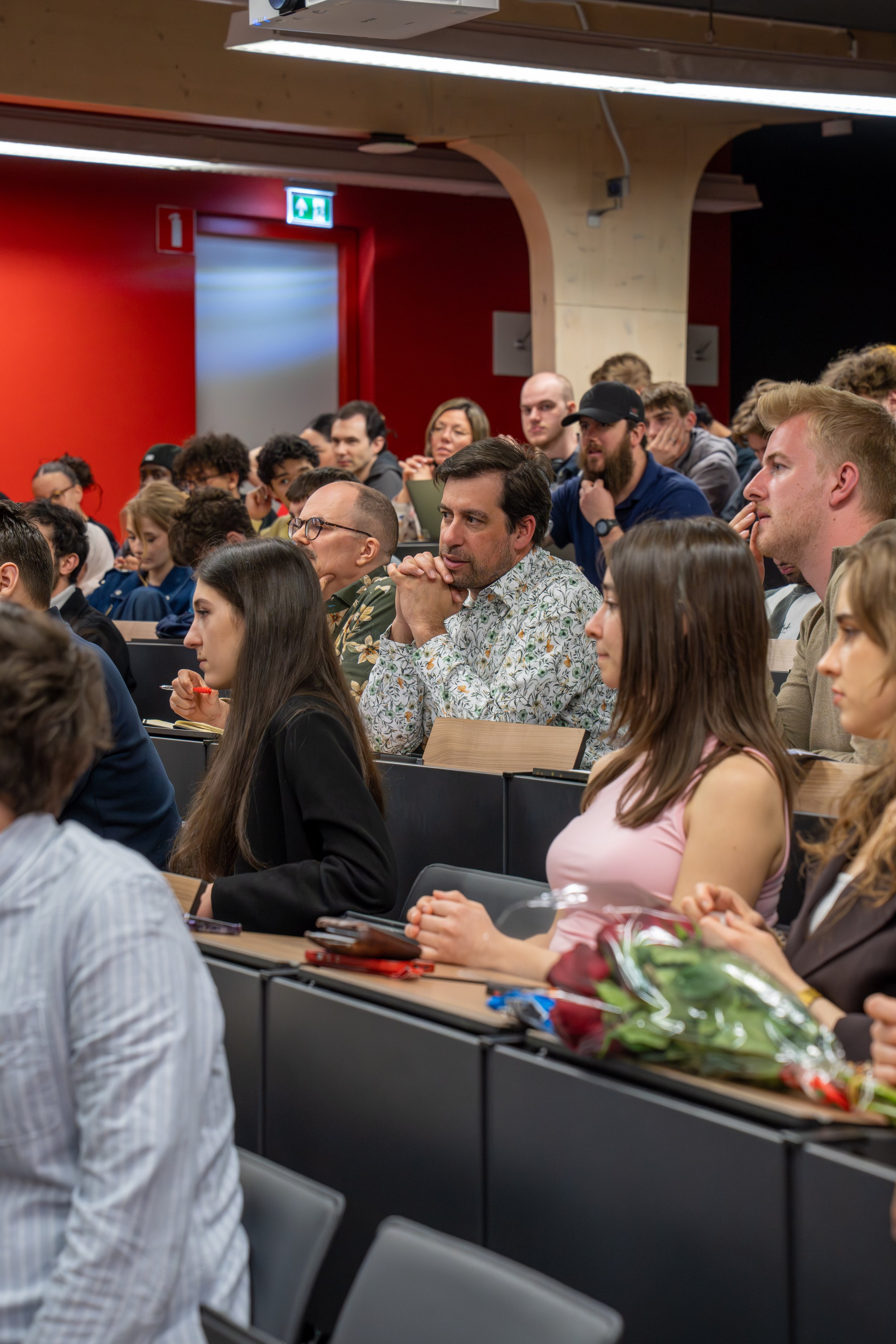 A large group of people seated at tables, attending a presentation or conference in a room with red walls and wooden accents. There are flowers on the table in the foreground.