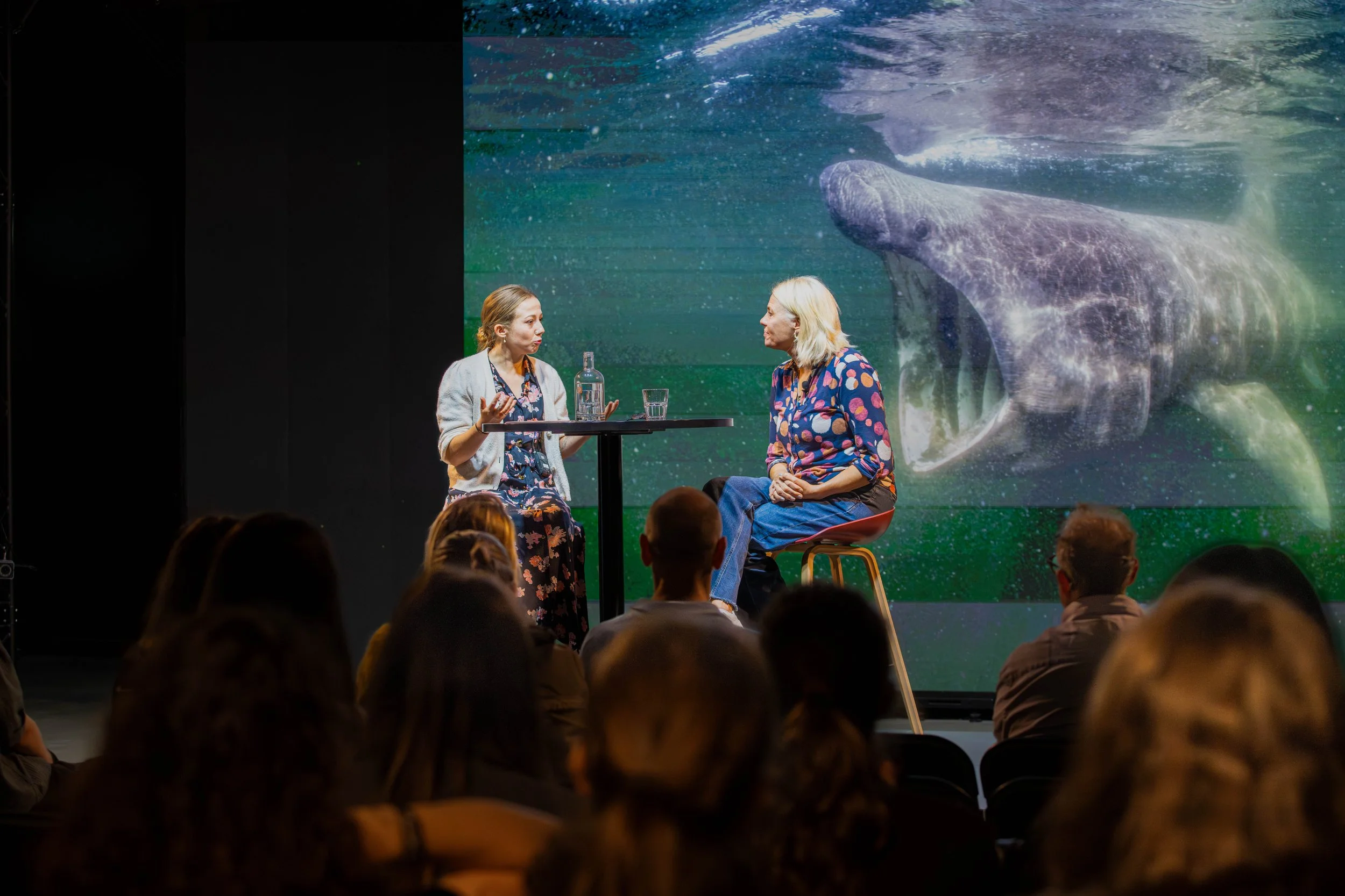 Two women sitting at a small round table engaging in a conversation during a presentation or event. A large screen behind them displays an image of a whale underwater, creating an oceanic background. Audience members are visible in the foreground.