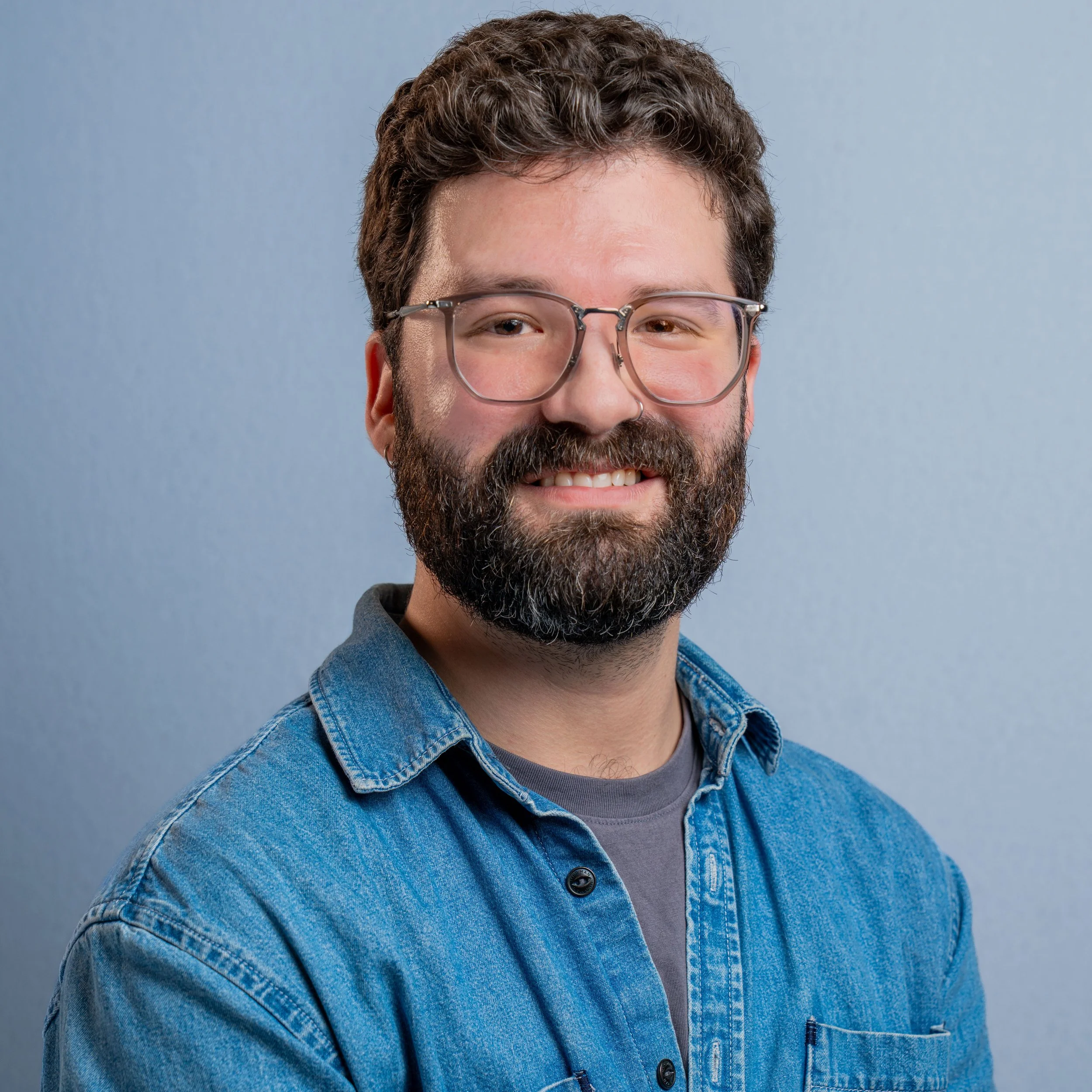 Close-up of a smiling man with curly brown hair, glasses, and a beard, wearing a denim jacket and grey shirt against a light blue background.