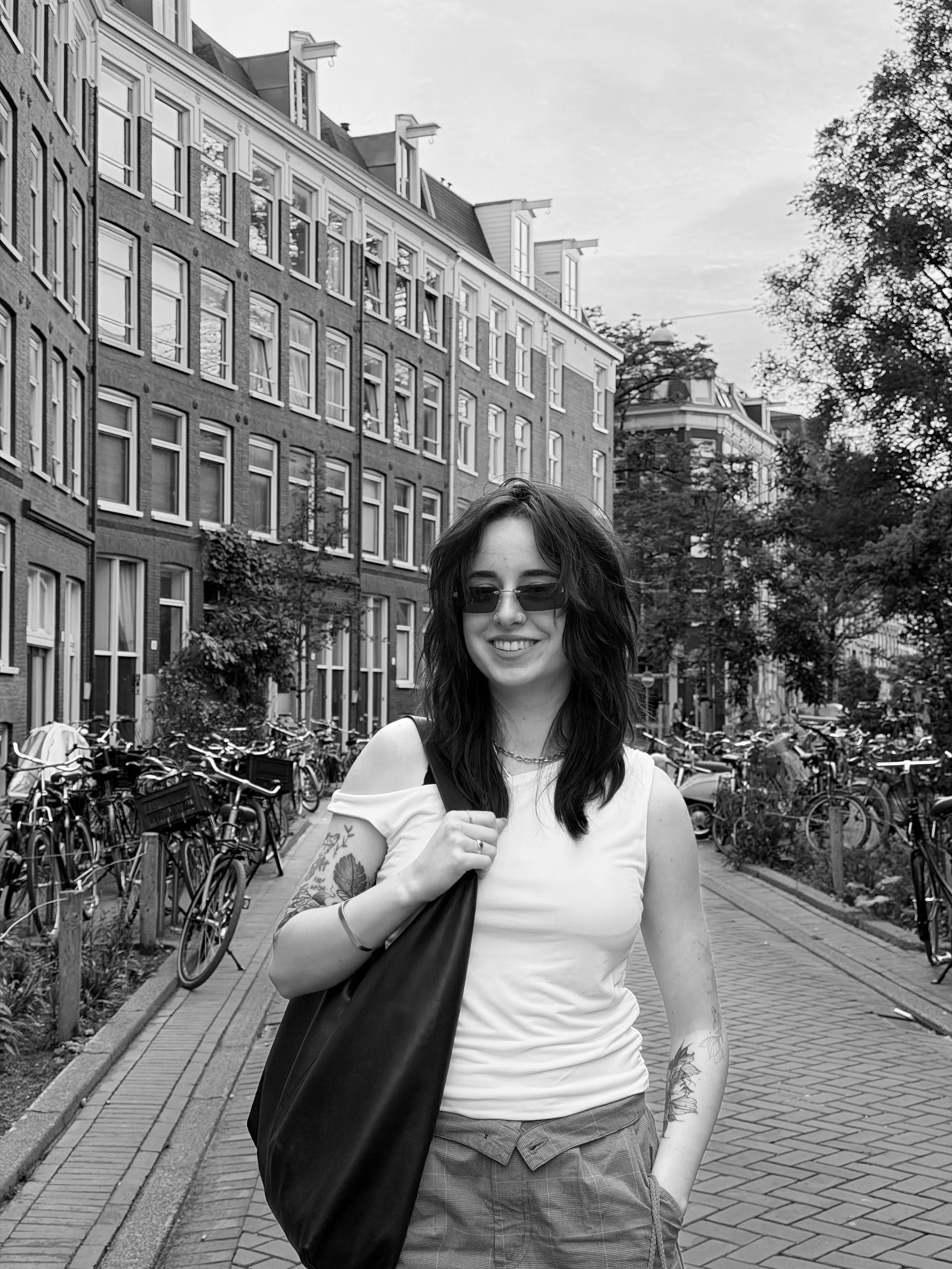 A young woman with dark wavy hair, wearing sunglasses, a white off-shoulder top, and a plaid skirt, walking on a city street with bicycles parked on the side, trees, and old European-style residential buildings in the background.