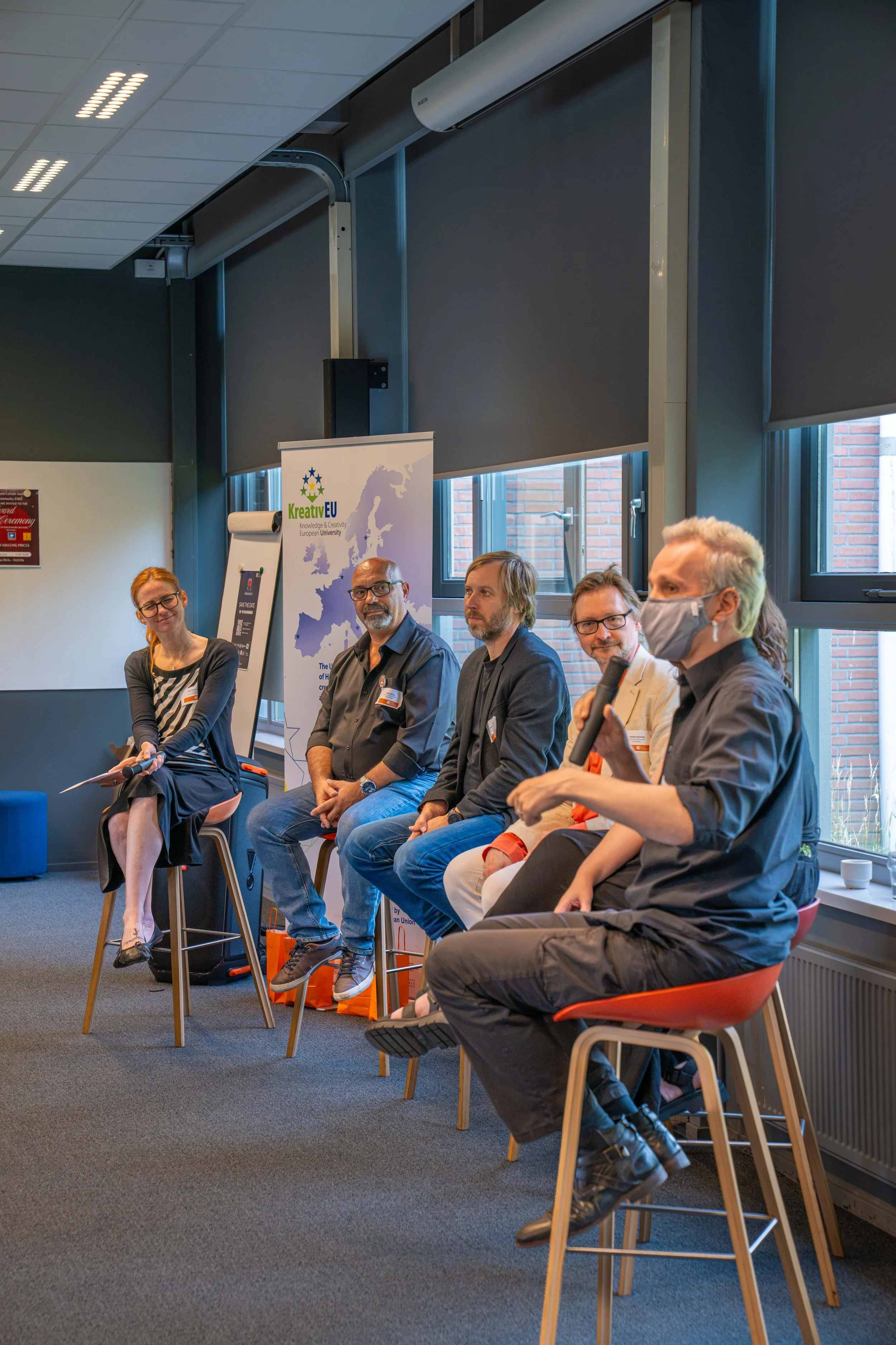 A group of five people sitting on chairs in a room with large windows. They are participating in a discussion or panel. There is a woman at the left holding a microphone, and a man at the right wearing a face mask. A banner with the logo and name 'Kreativ EU' is visible in the background.