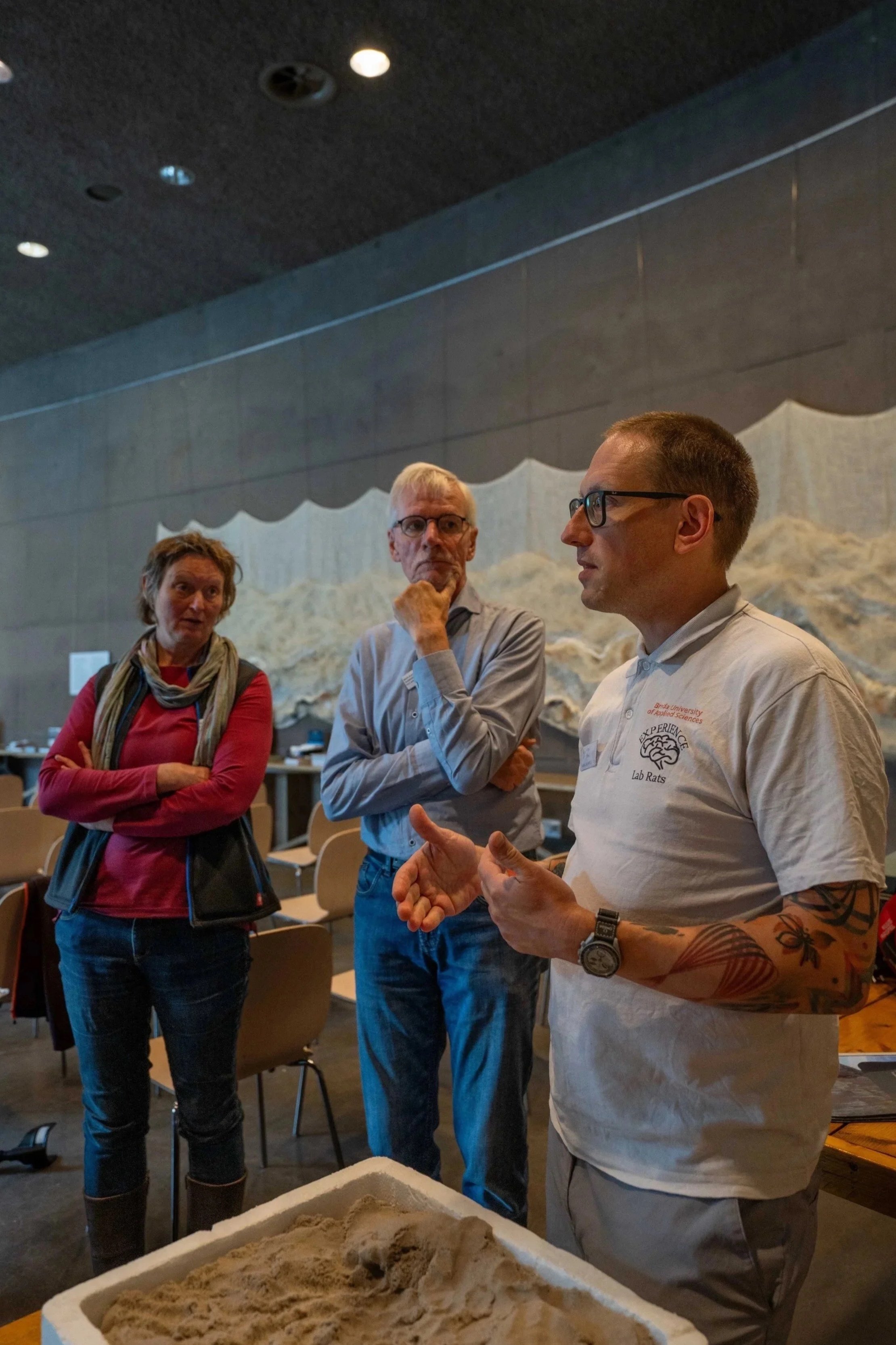 Three people engaged in conversation indoors, with one man gesturing as he talks. A woman with a scarf and a man with glasses listen. In front is a large container of sand.