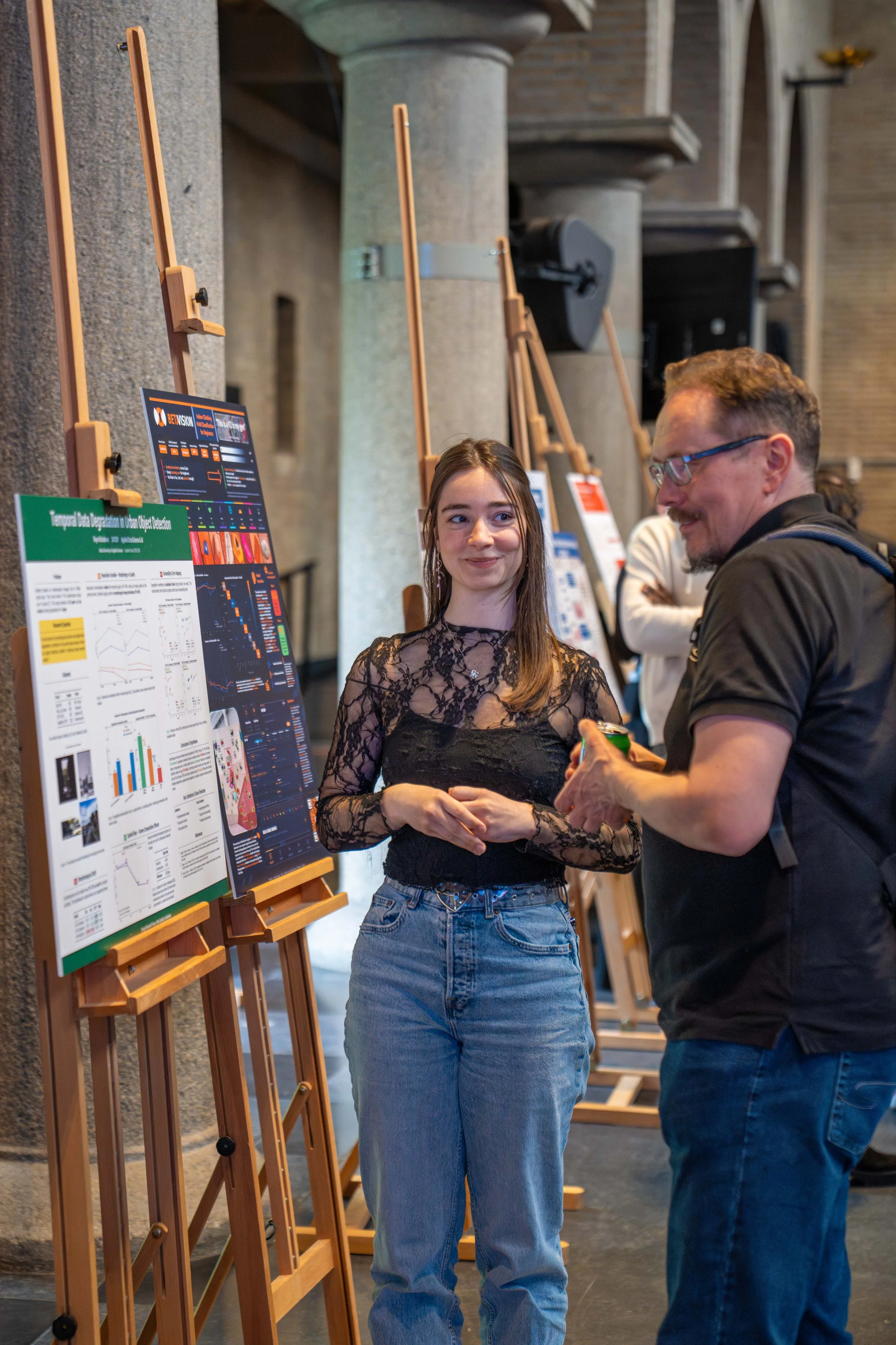 A young woman and an older man are standing in front of presentation posters at an indoor event. The woman has long hair and is wearing a black lace top and baggy jeans, while the man is wearing glasses and a dark shirt. They are engaged in conversation near informational displays on easels.