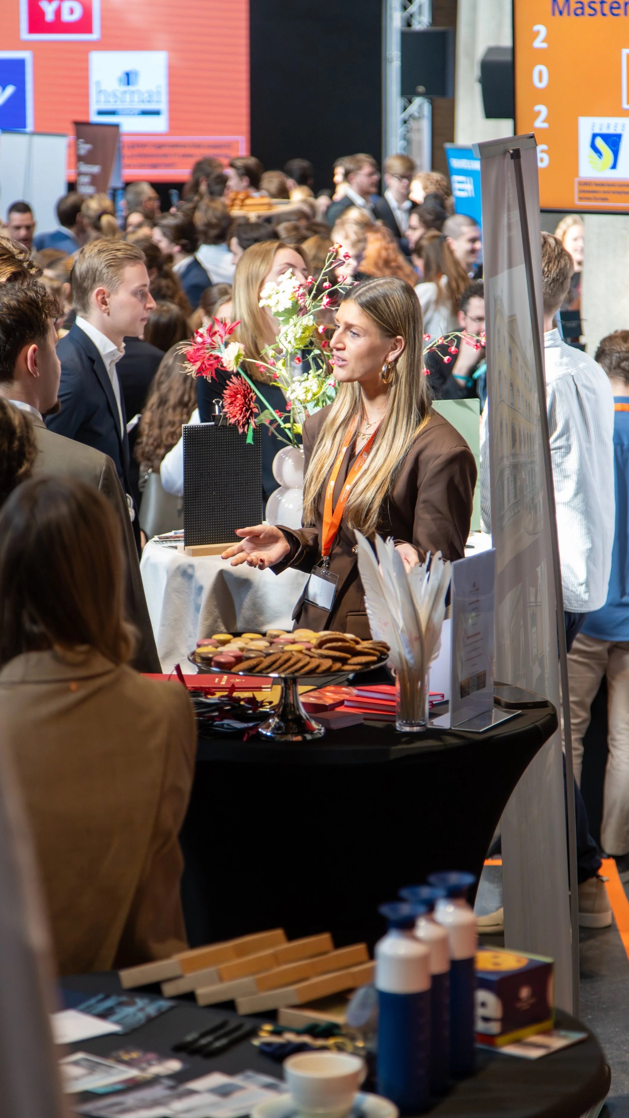 A crowded indoor event with a booth displaying cookies, brochures, and a large flower arrangement. People are engaged in conversations, and large screens with text are visible in the background.