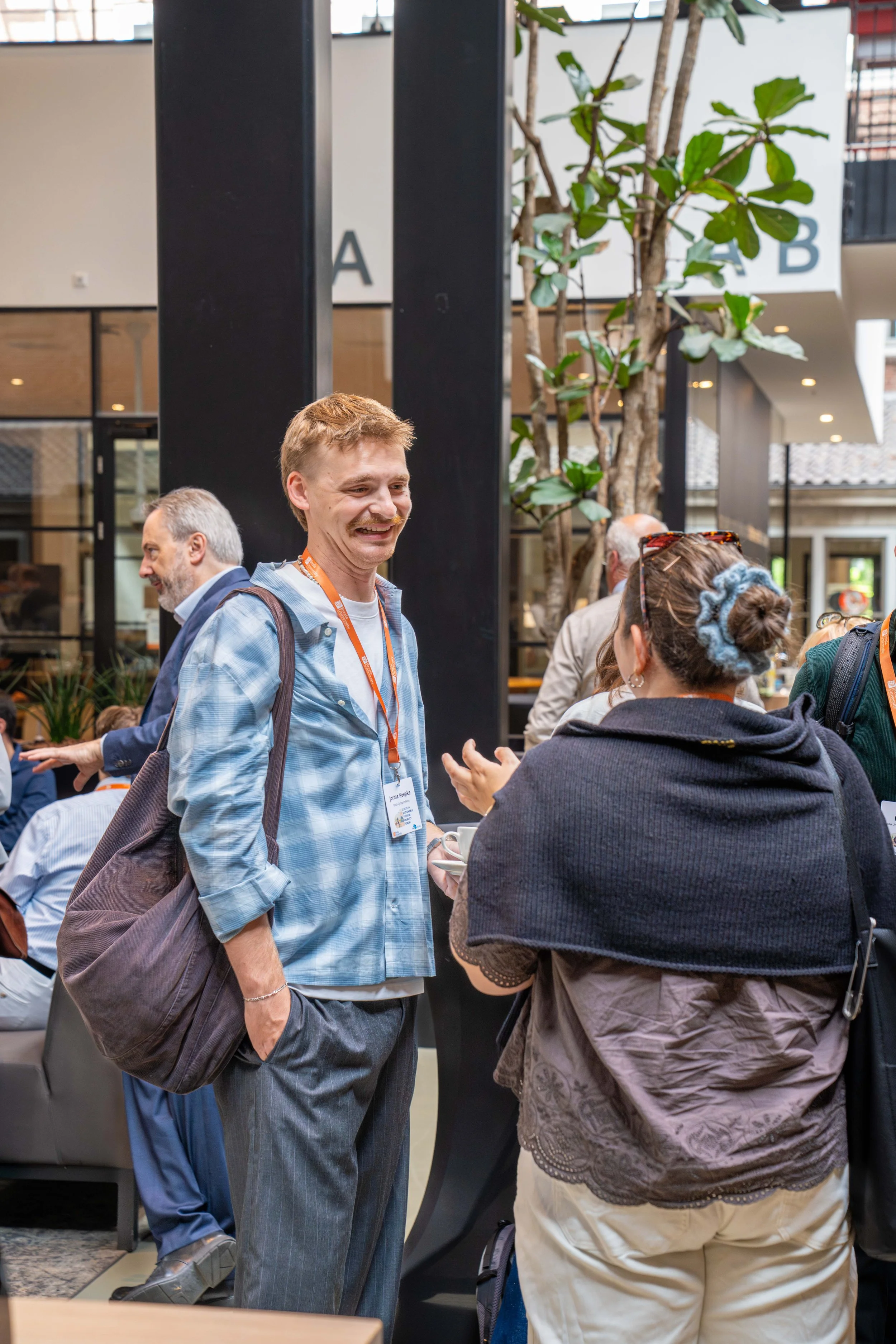 People socializing at an indoor event, with a man in a blue plaid shirt and gray pants talking to a woman in a dark sweater. There are other individuals in the background, and signs labeled 'A' and 'B' are visible above a tall tree inside the venue.