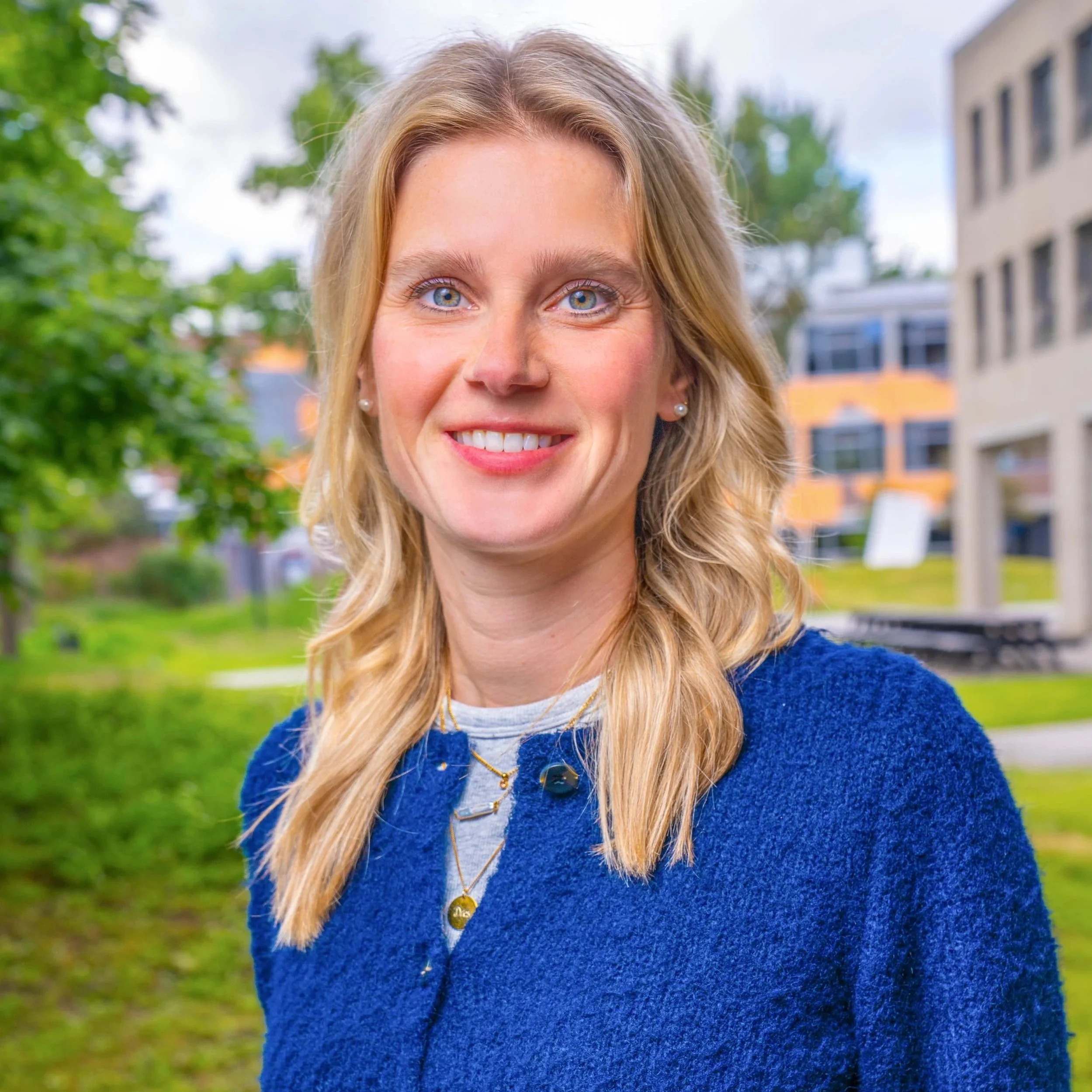Close-up of a woman with blonde hair, wearing a blue sweater, smiling outdoors with trees and buildings in the background.