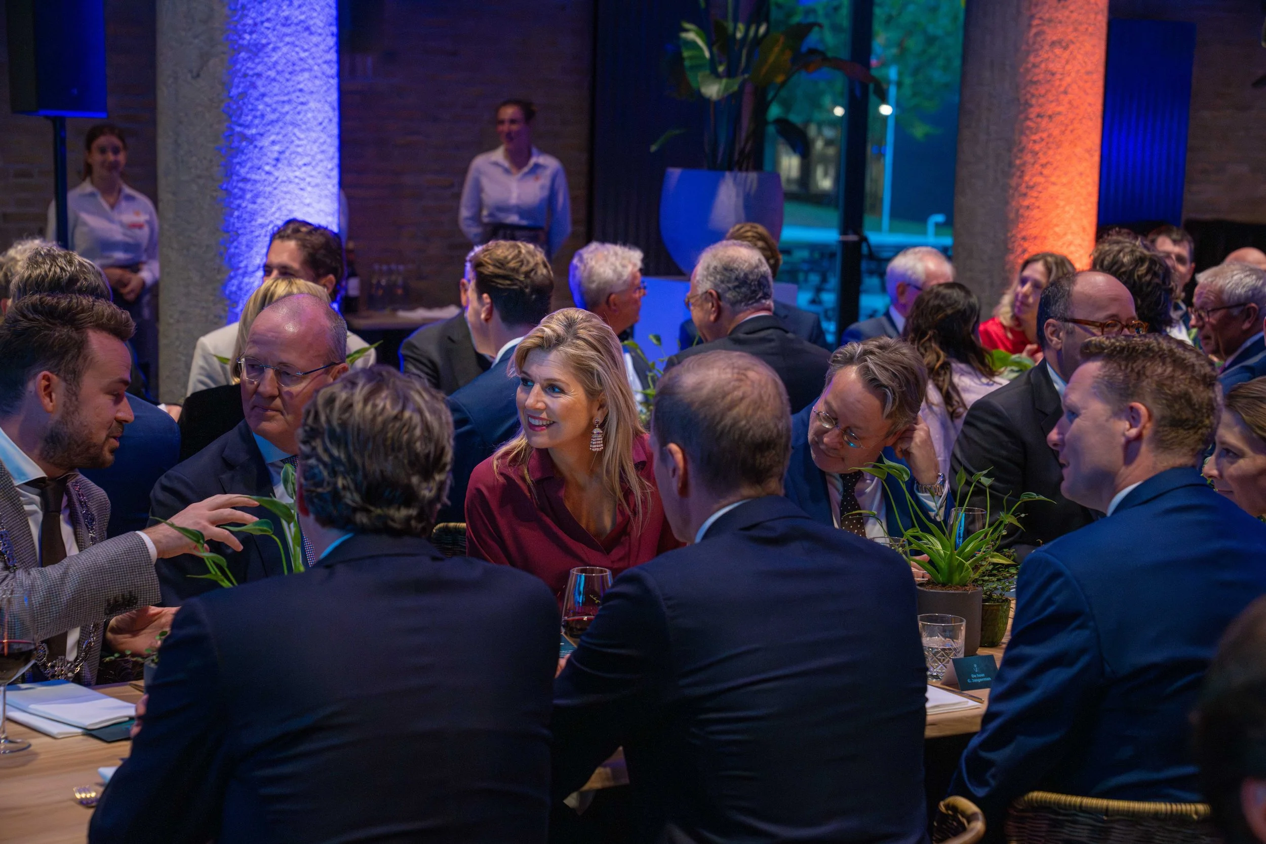 People seated at a long table in a dimly lit room, attending an event or conference, with some individuals engaged in conversation and others smiling, with a large decorative plant and background lighting adding to the ambiance.