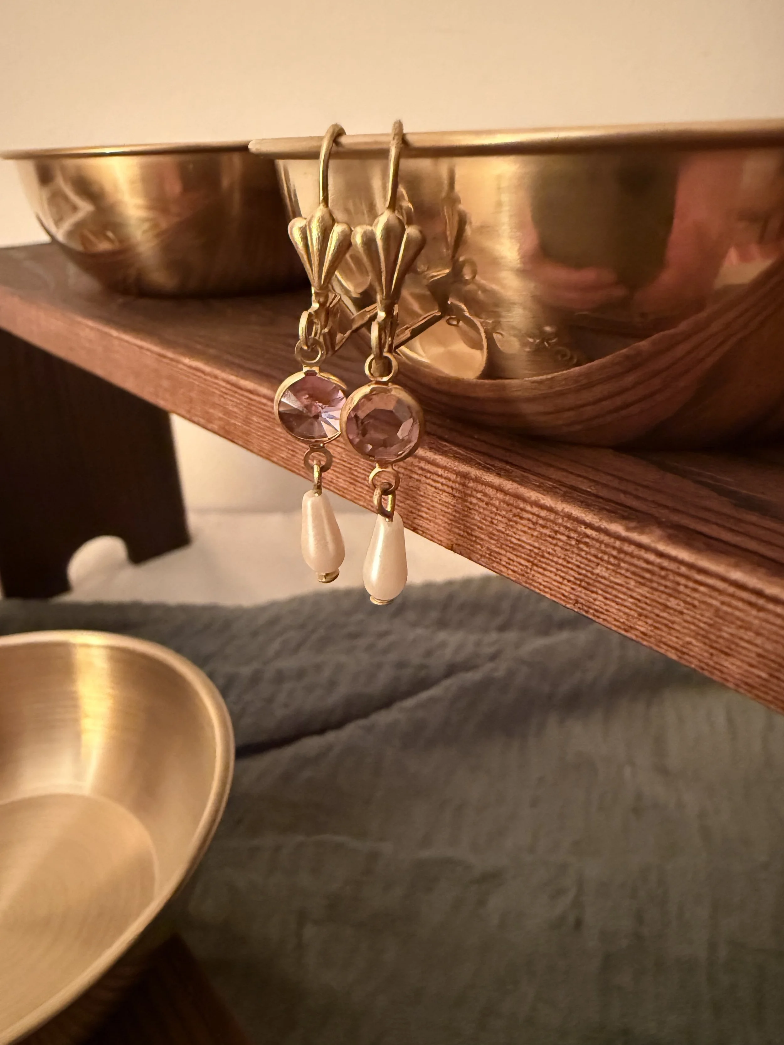 Close-up of a pair of gold earrings with large faceted pink stones and white teardrop-shaped pendants hanging, displayed on the edge of a brass bowl on a wooden surface.