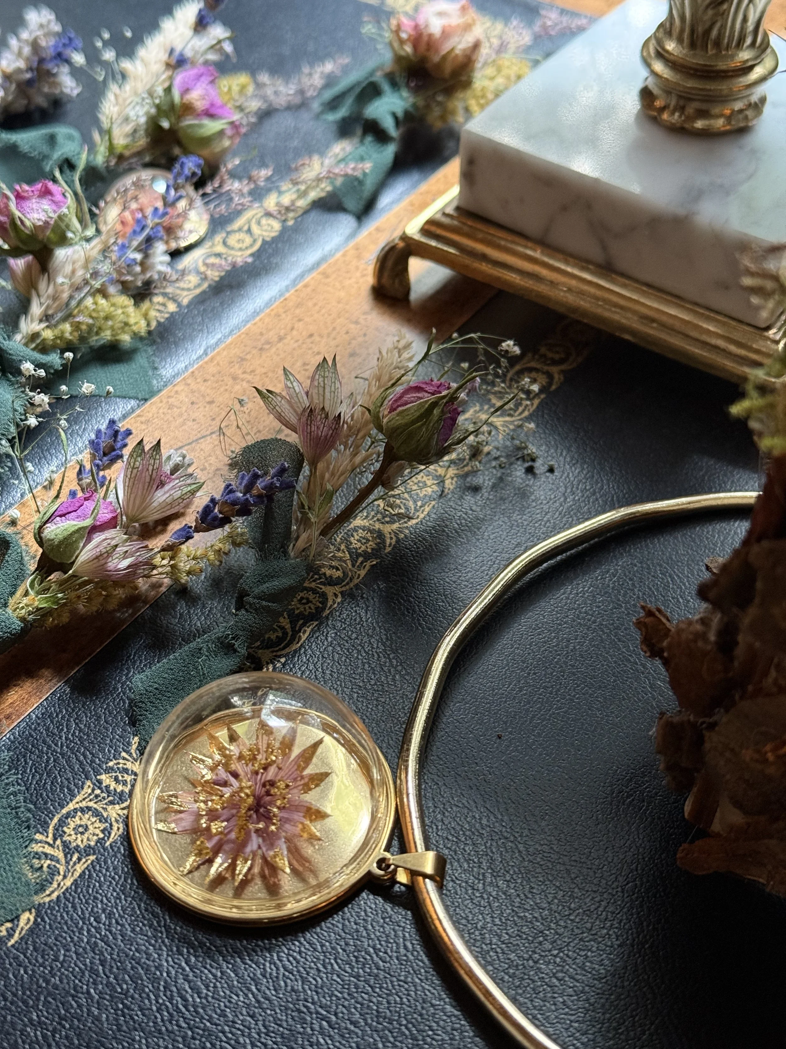 Close-up of dried pressed flowers arranged on a decorative cloth, with a jewelry magnifying glass, ornate jewelry box, and a piece of carved wooden furniture in the background.