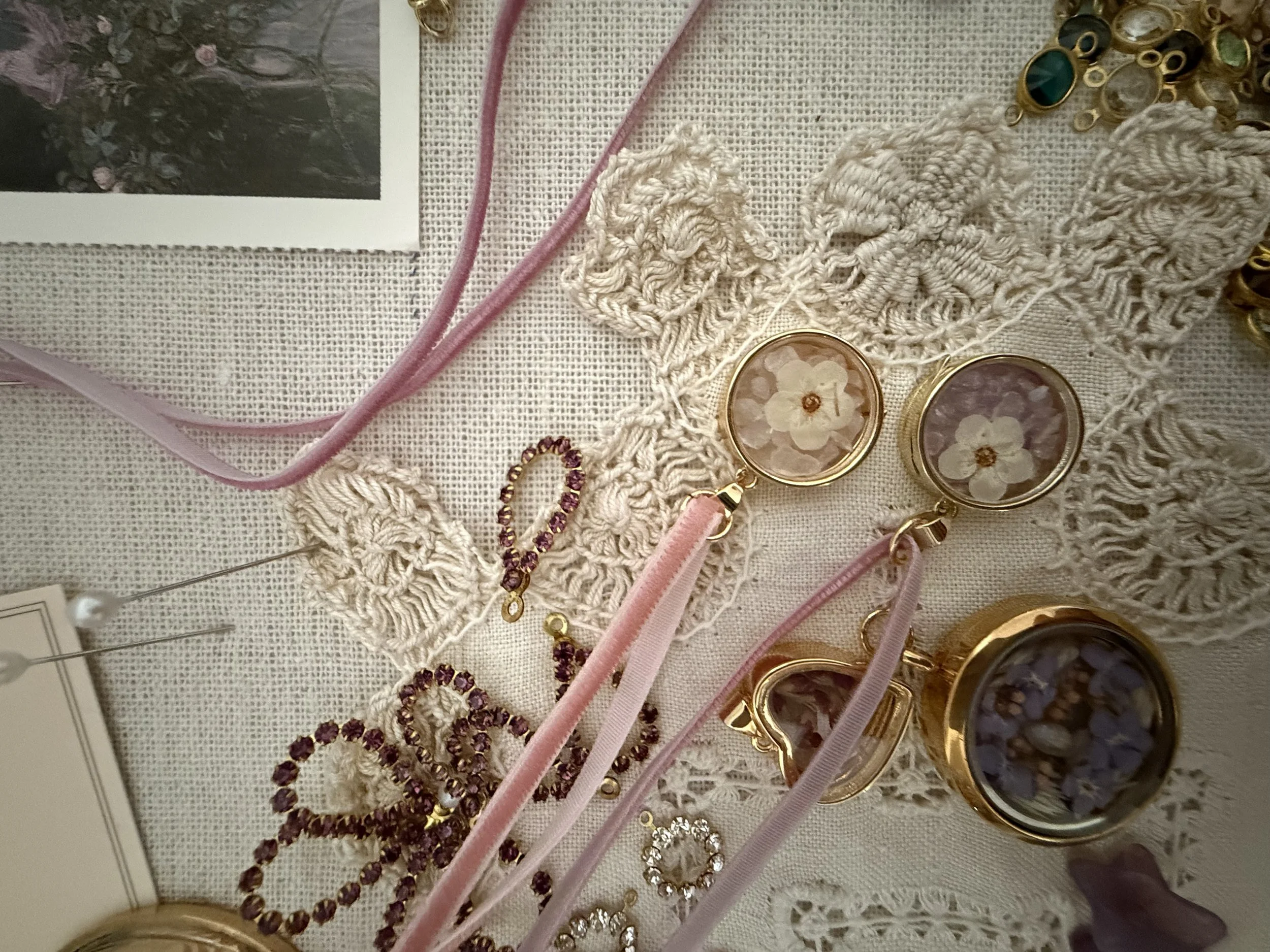 Assorted jewelry and accessories, including floral-patterned sunglasses, pink velvet ribbon, crocheted lace, and various rings and bracelets, arranged on a textured fabric surface.