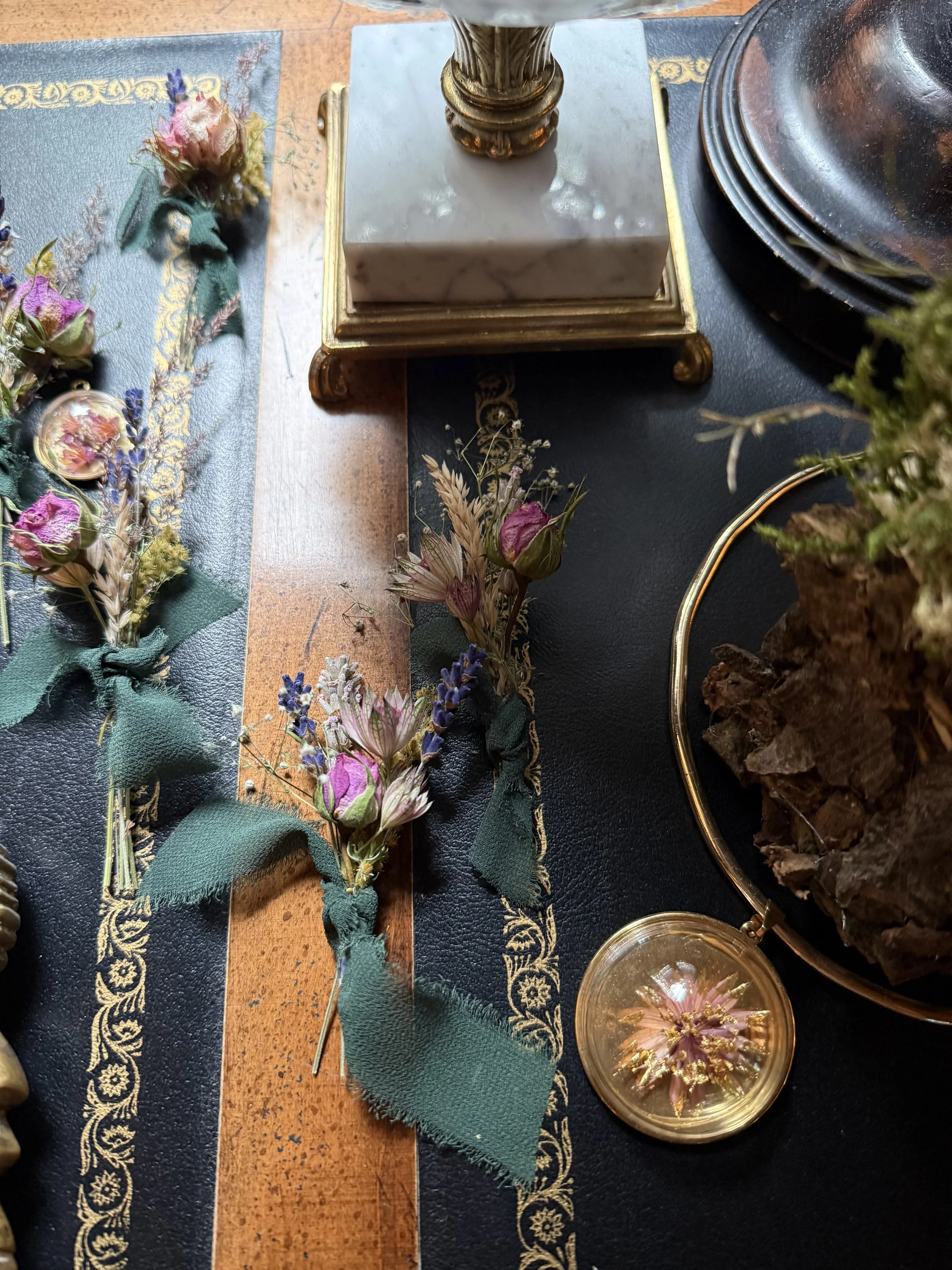Arrangement of dried flowers and plants on a table, with decorative items including a small marble and gold stand, plates, and a glass container, all on a dark and wooden surface.