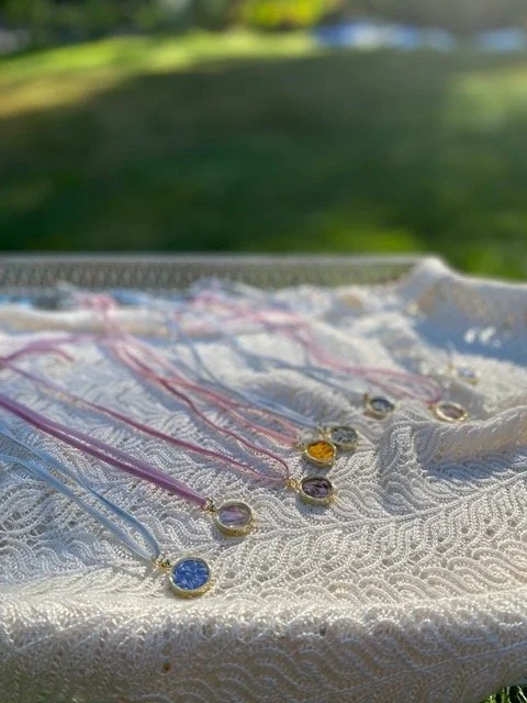Collection of colorful necklaces with round pendants laid out on a white textured cloth outdoors with green grass and trees in the background.