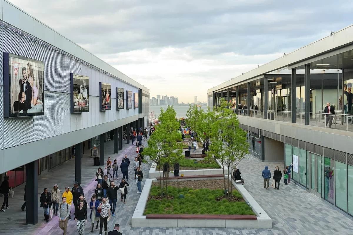 Outdoor shopping mall with people walking along a paved walkway between two modern buildings. The left building has advertisements on the windows, and the right building has large glass windows. There are trees and green planters in the center of the walkway, with the city skyline visible in the background across a body of water.