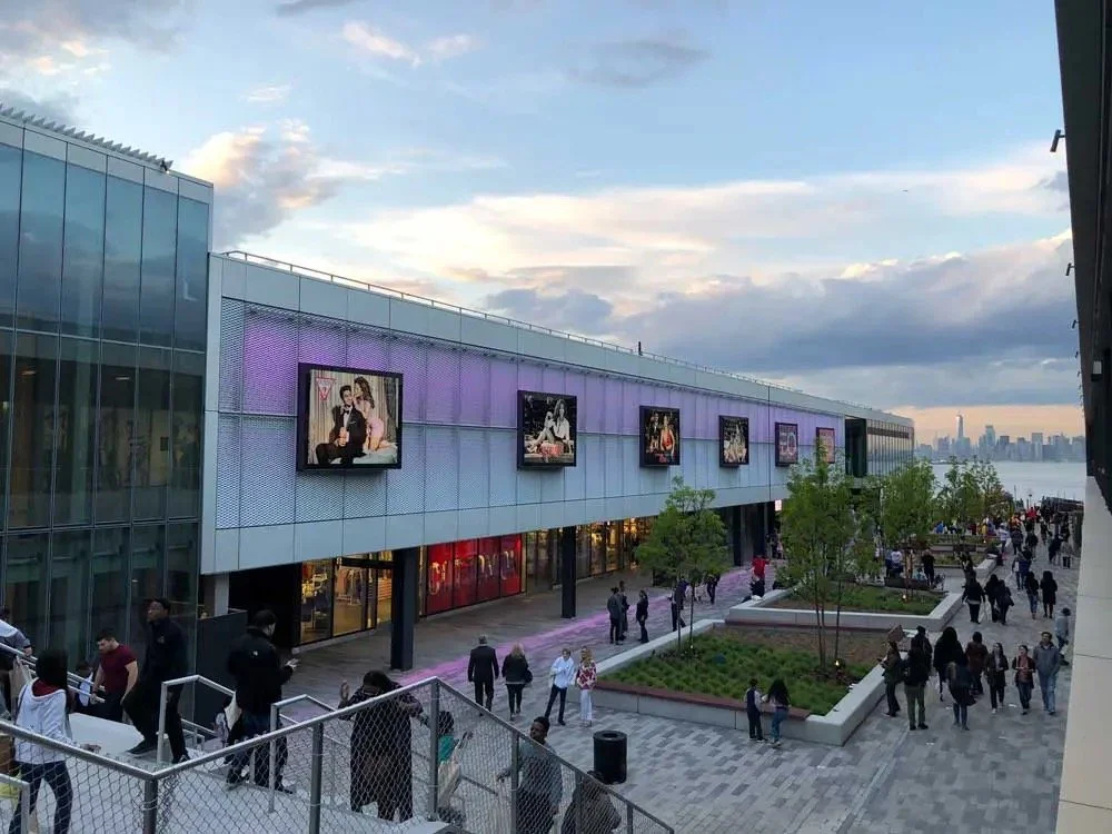 Outdoor shopping mall with a modern glass building, digital billboards displaying fashion ads, and people walking along a paved promenade with benches and small trees, overlooking a city skyline in the distance.