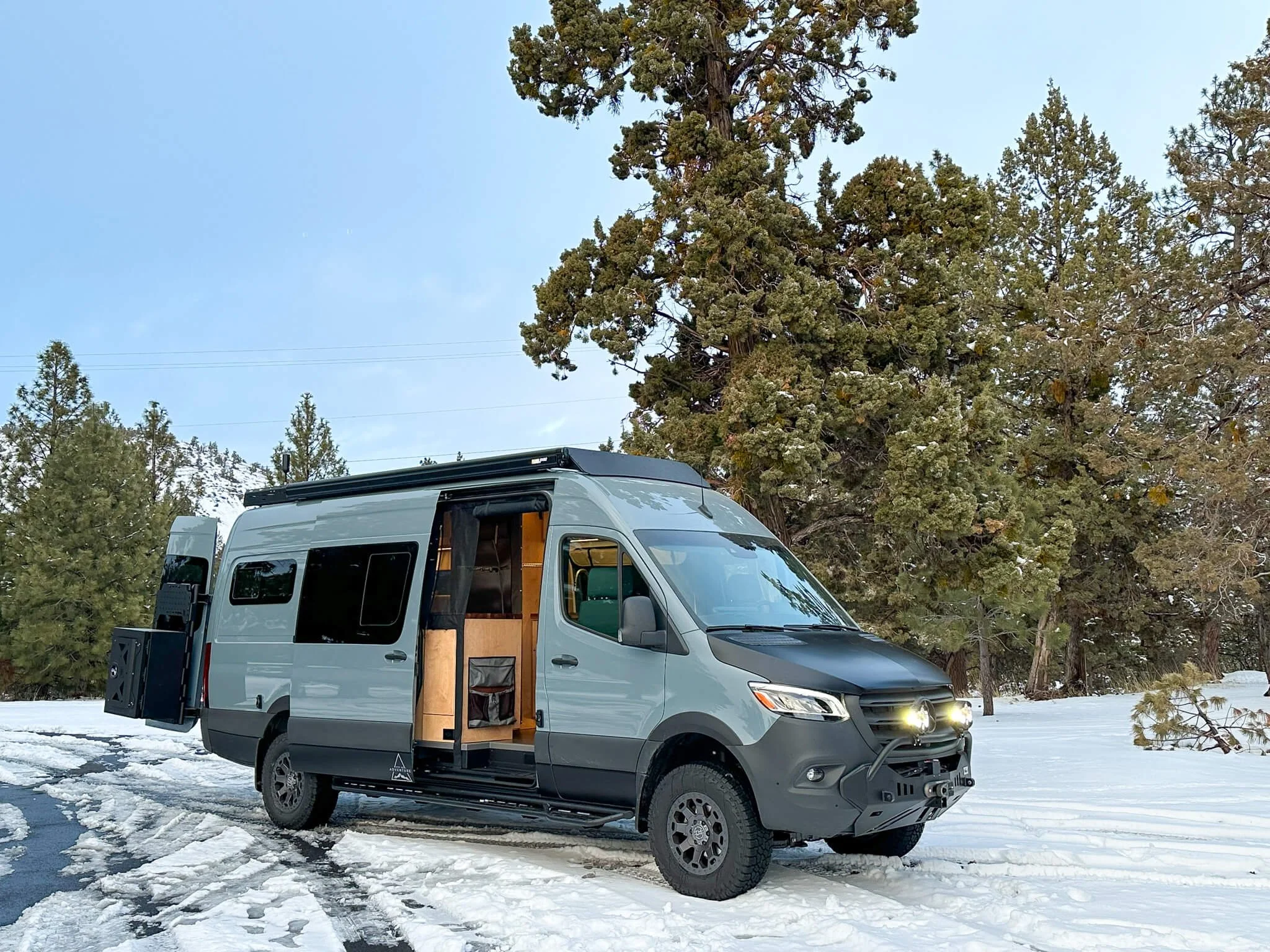 Open slider door revealing custom HWA interior of 'Dusty Roads' 170 Sprinter van in snowy setting.